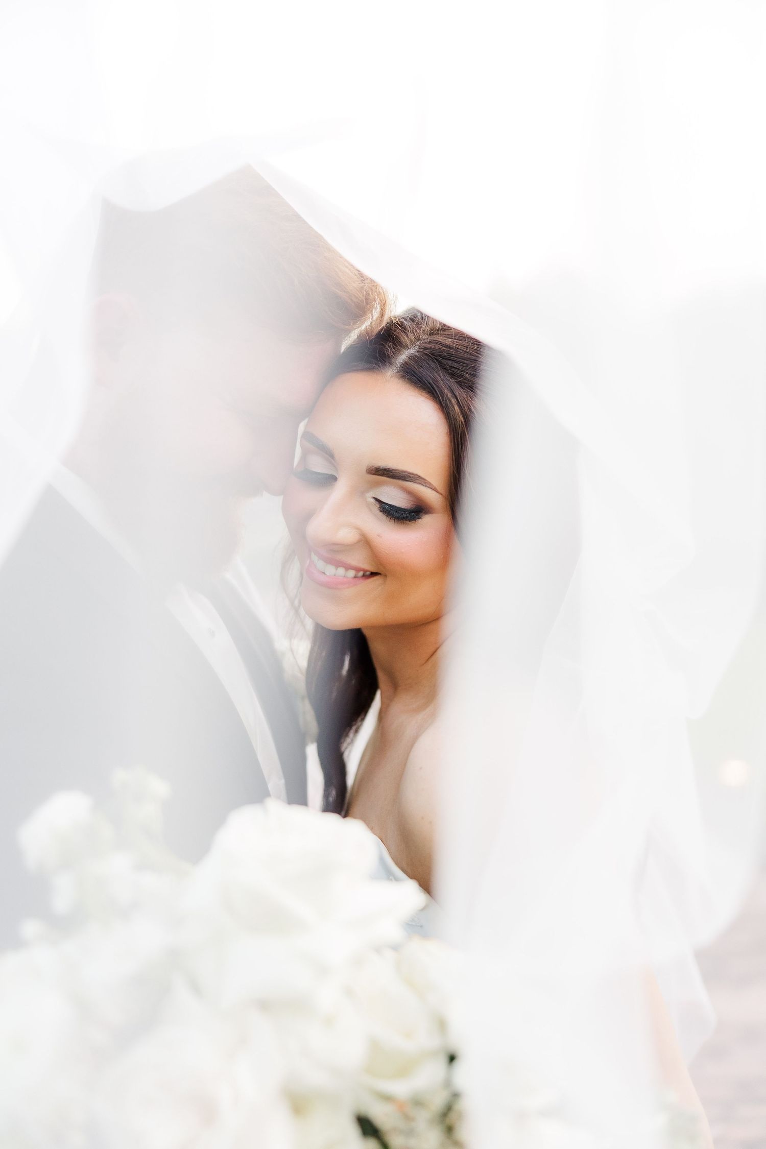 Artistic bridal portrait through sheer veil with soft lighting and white flowers.