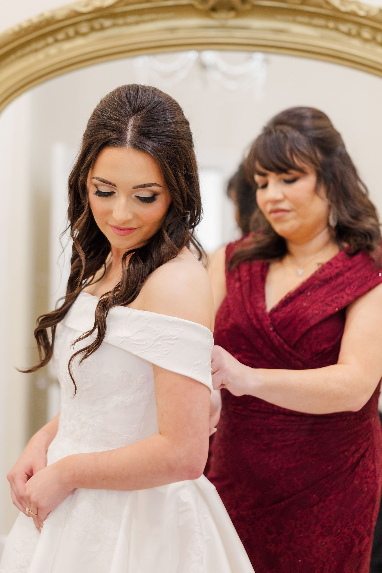 Two people preparing for a wedding, with one wearing a white off-shoulder dress being helped by another in burgundy.