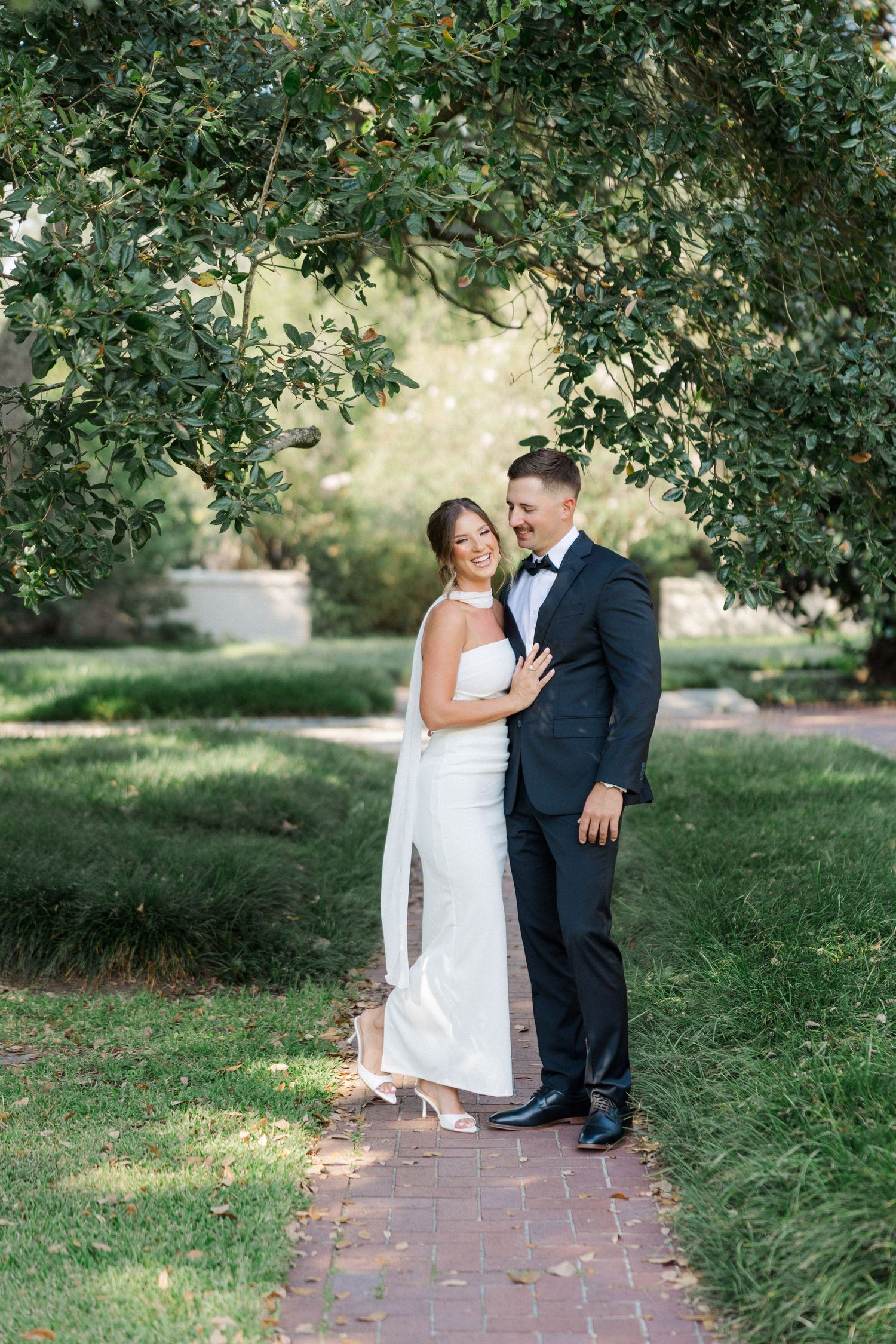 A couple in formal wedding attire pose under a lush green tree archway on a brick path in a garden setting.