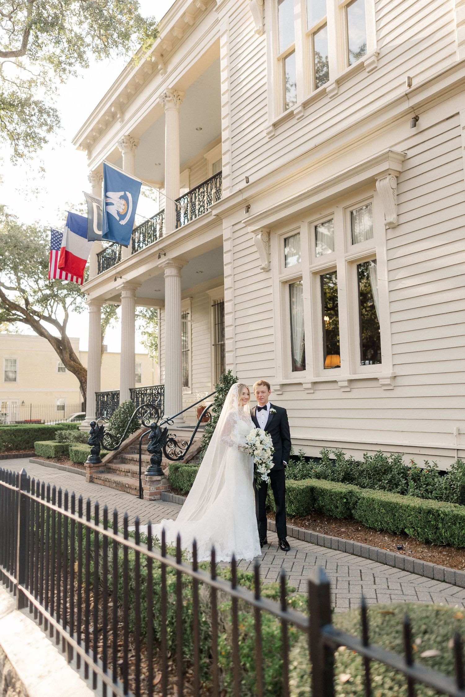 Wedding couple poses outside a grand white colonial building with flags and iron fencing.