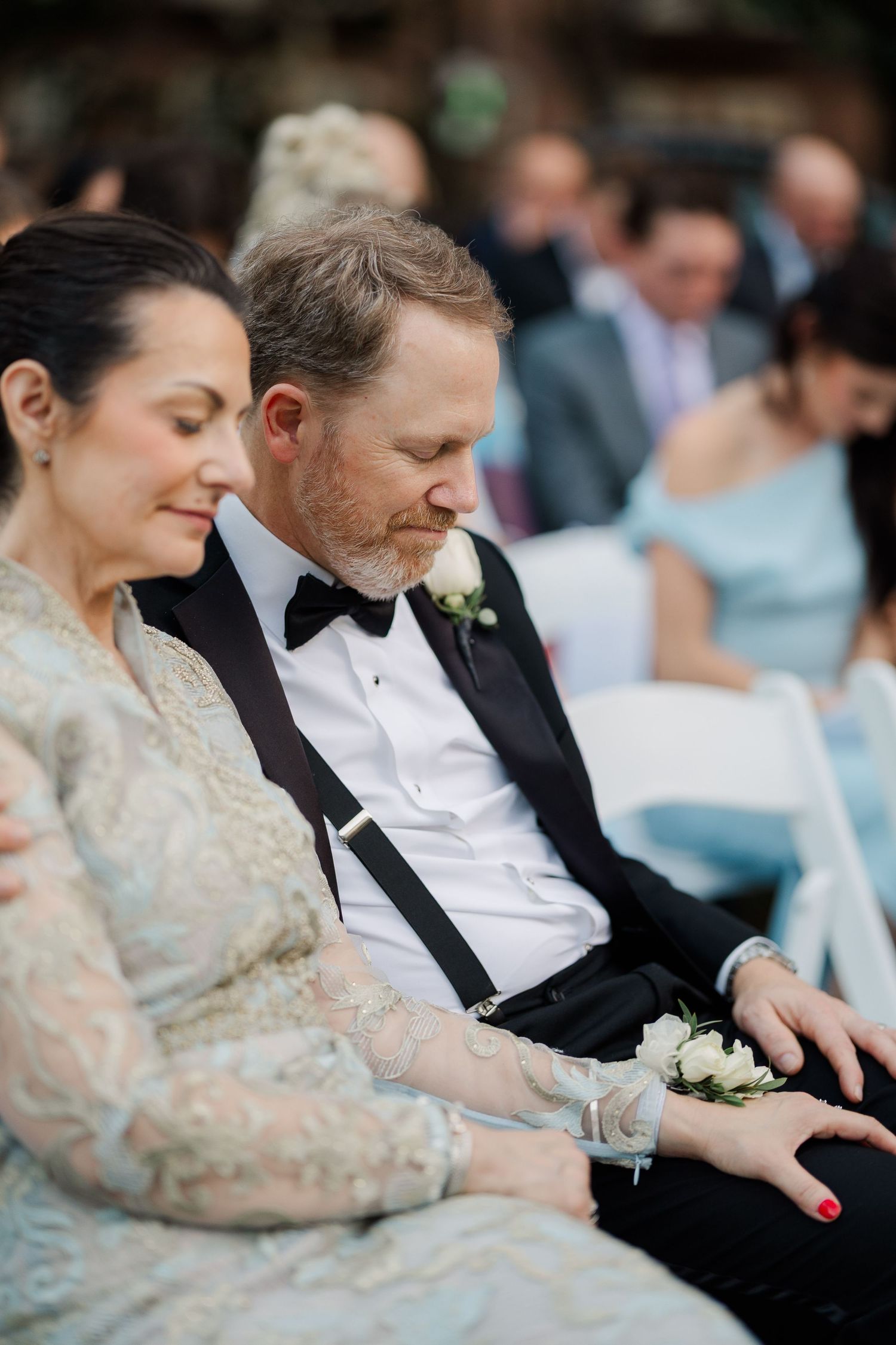 Couple in formal attire sitting together at outdoor wedding ceremony with guests in background.