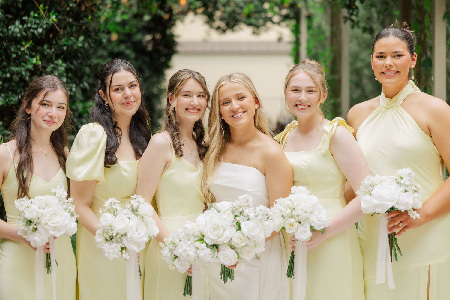 Row of bridesmaids in pale yellow dresses holding white bouquets stand together at outdoor wedding ceremony.
