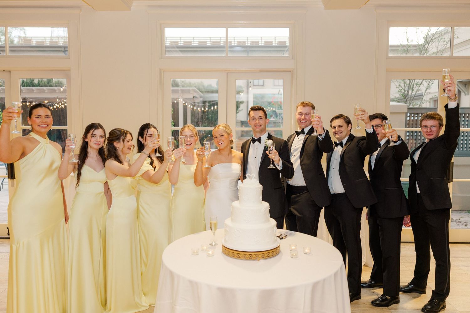 Wedding party celebrates around white cake with bridesmaids in yellow and groomsmen in black.