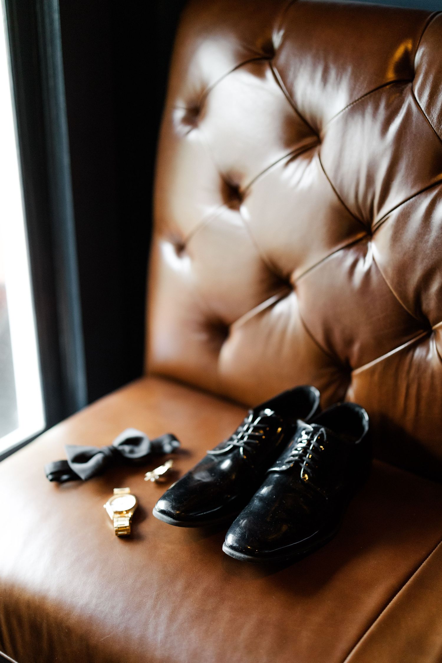 A pair of black leather dress shoes rests on a brown tufted leather couch or chair.