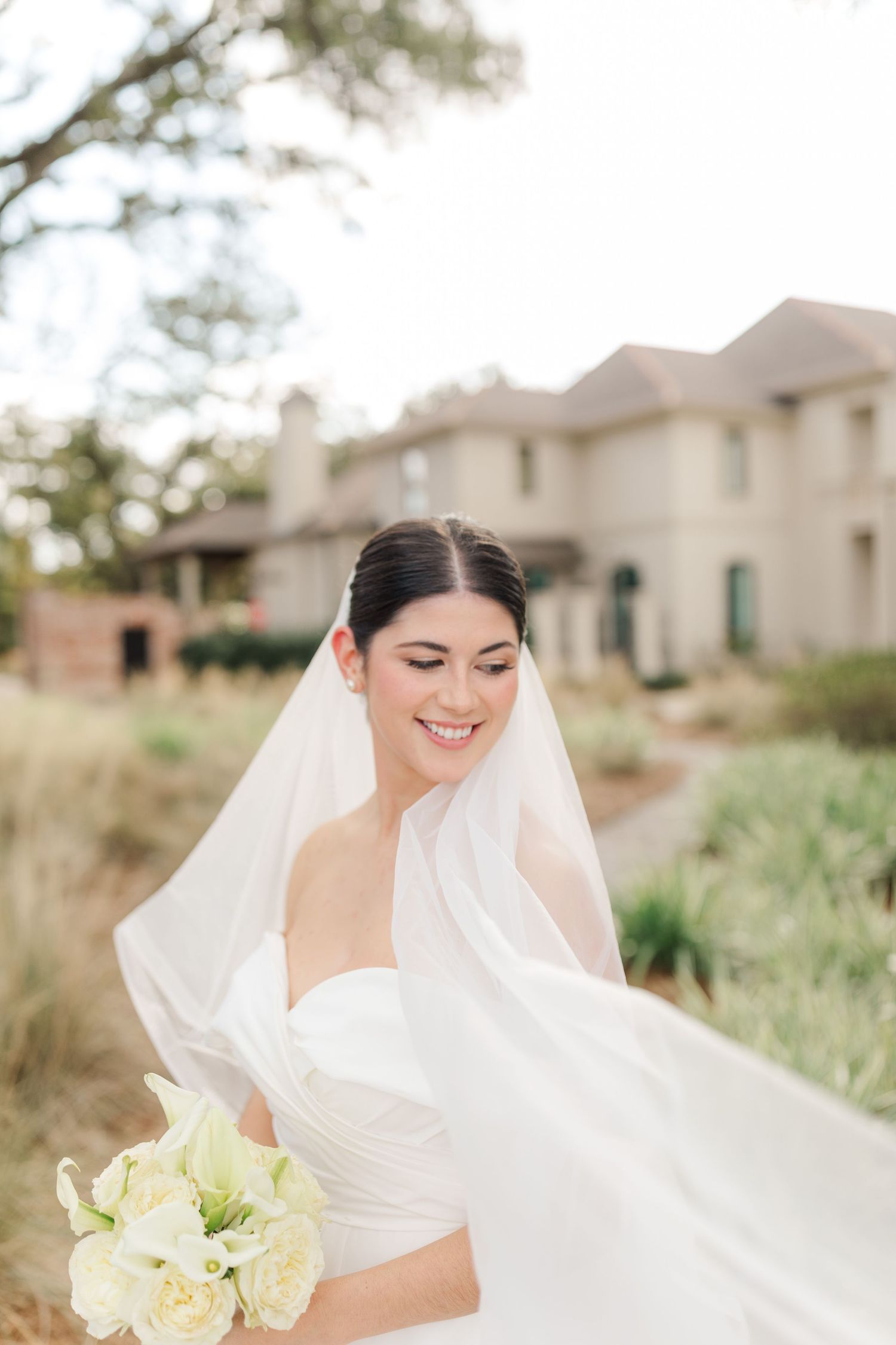 A bride in a white wedding dress with a flowing veil holds a bouquet of cream roses against an elegant building backdrop.