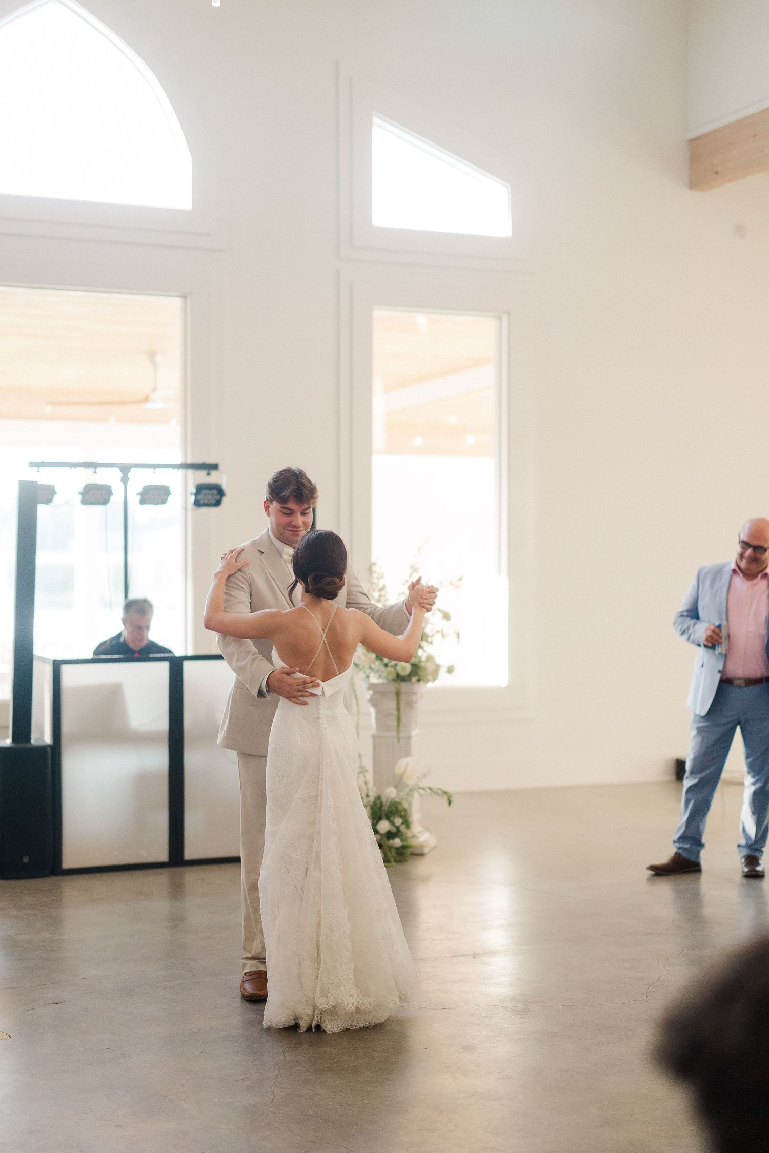 Bride and groom share first dance in bright white venue with arched windows and guests watching.