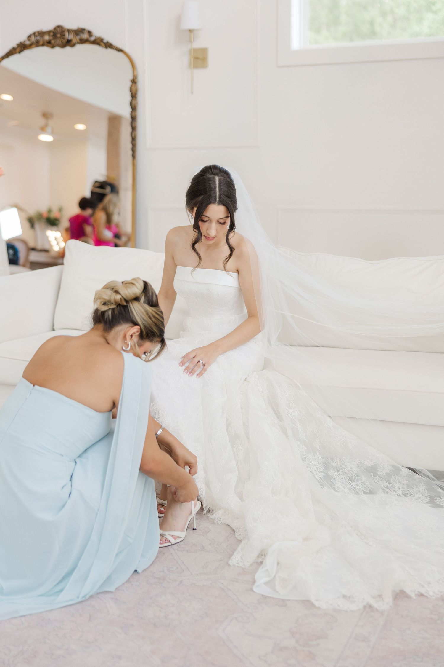Bridesmaid in light blue gown helping bride with shoes while seated on white sofa in bridal suite.