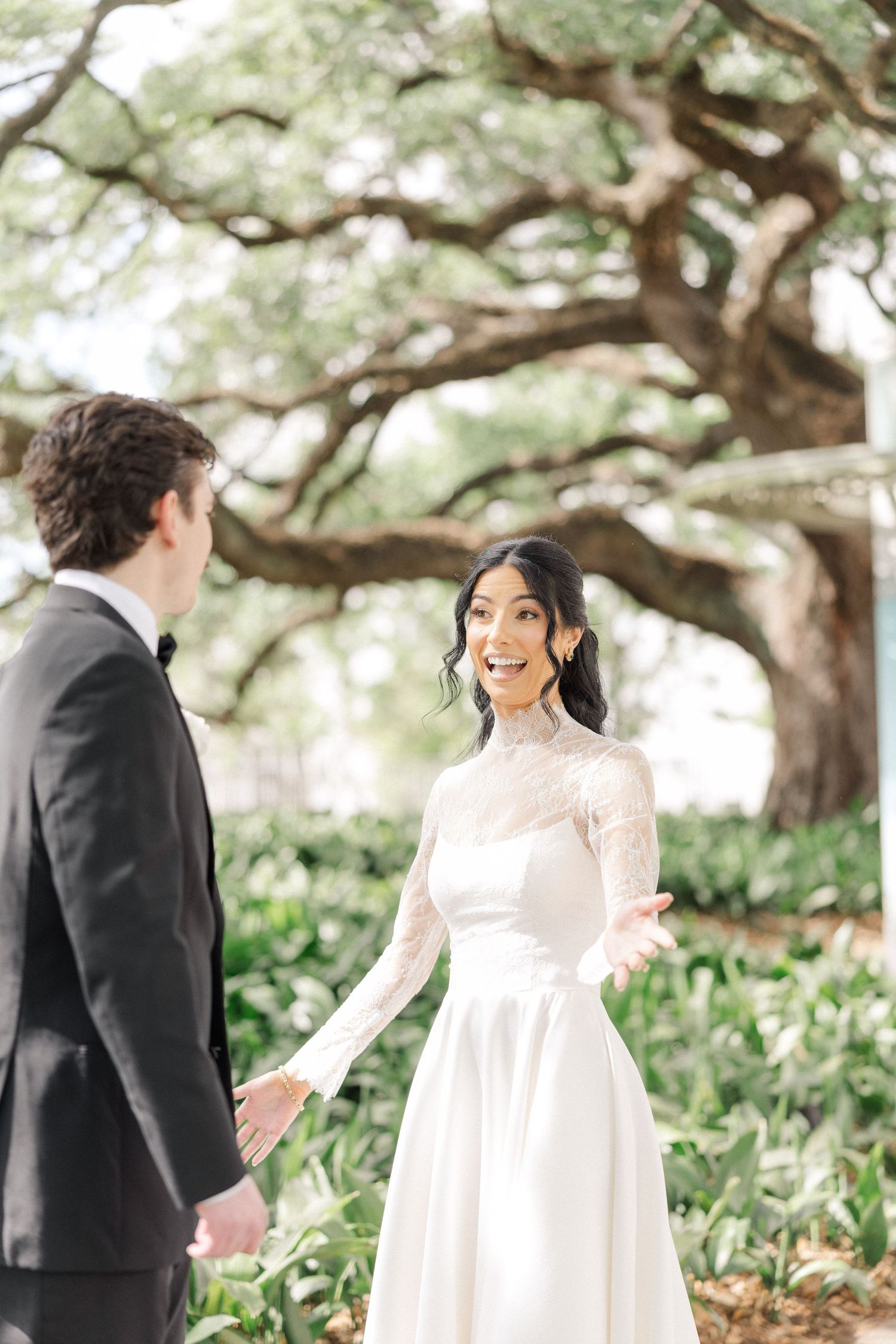 Bride in lace long-sleeve wedding gown reacts with joy during first look with groom under large oak tree.