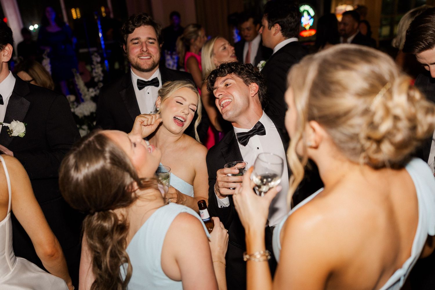 Wedding guests in formal attire laughing and dancing together at lively reception celebration.