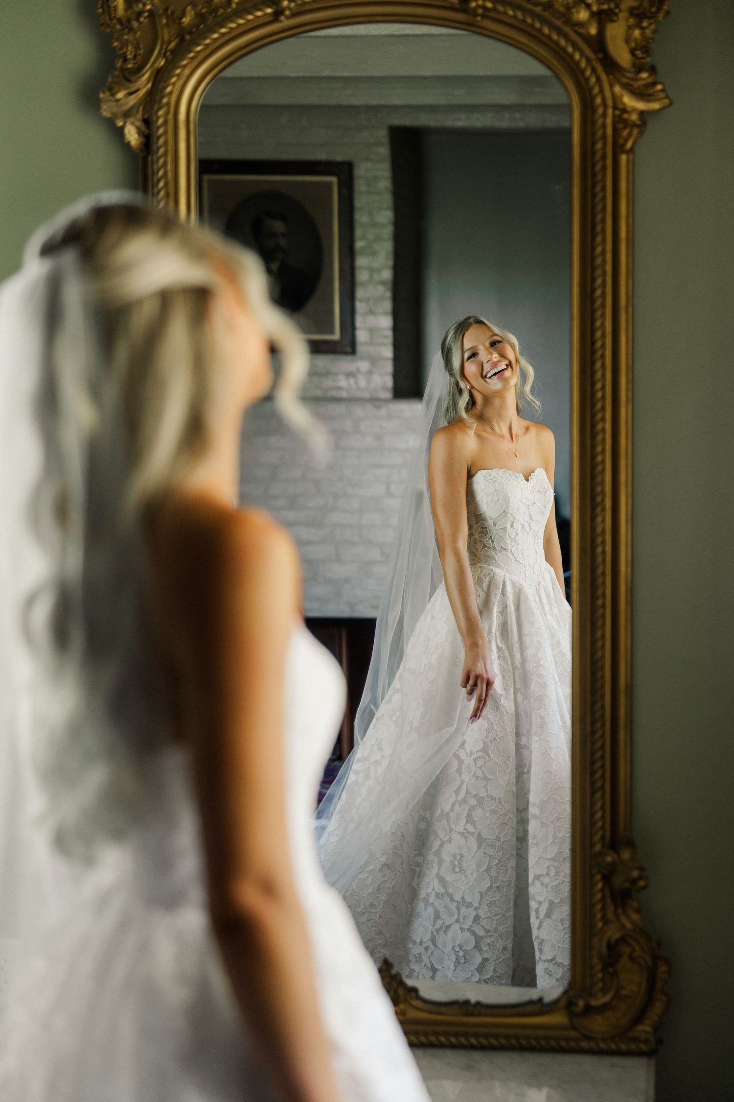 Blonde bride in strapless lace gown and veil smiling at her reflection in an ornate gold mirror.