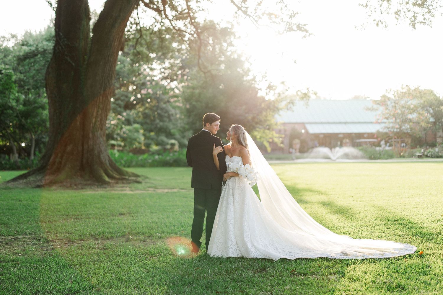 Bride and groom sharing a tender moment on sunlit green lawn with golden hour light and fountain backdrop.