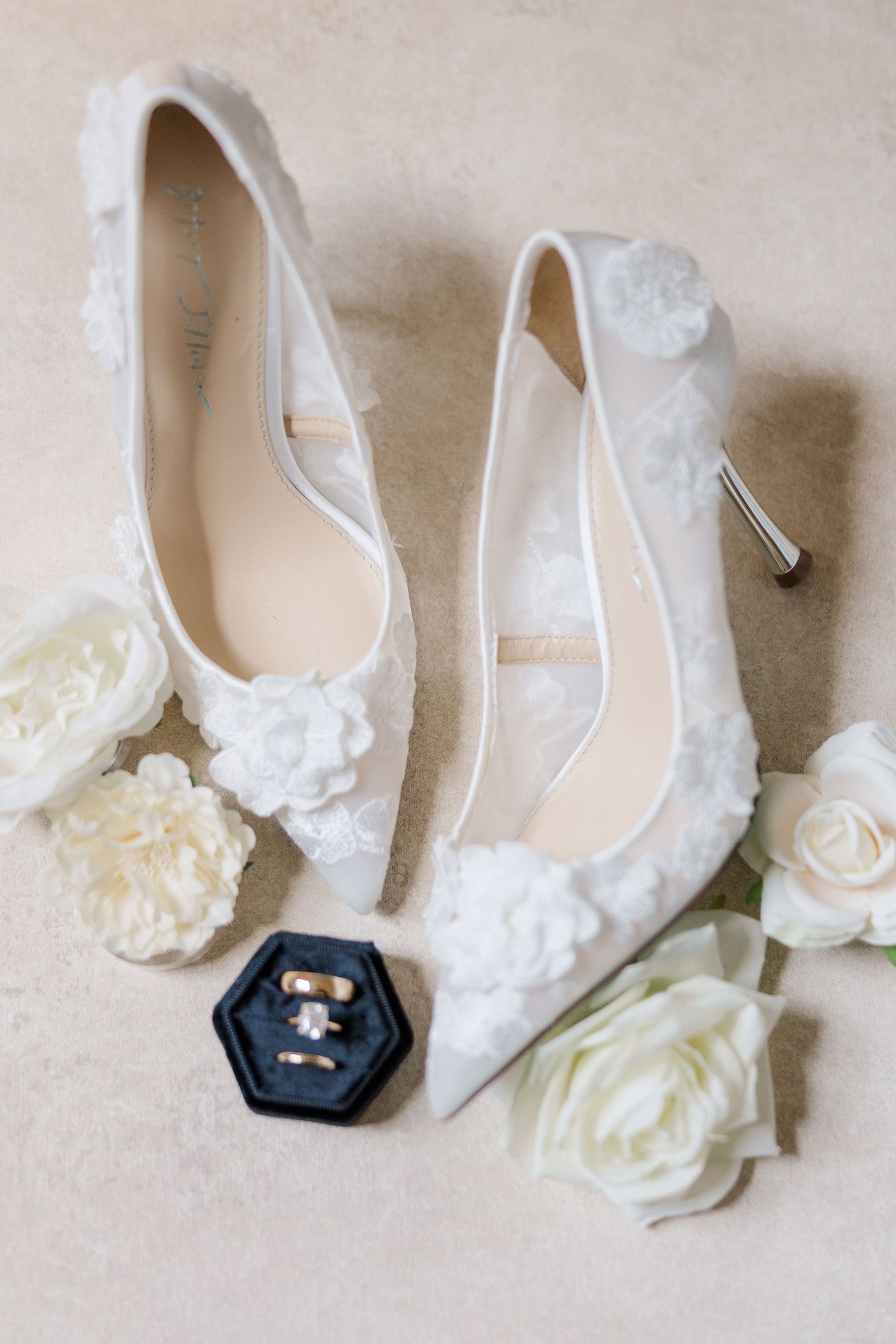 Elegant white lace wedding shoes with delicate floral details and a navy hexagonal ring box displayed on beige background.