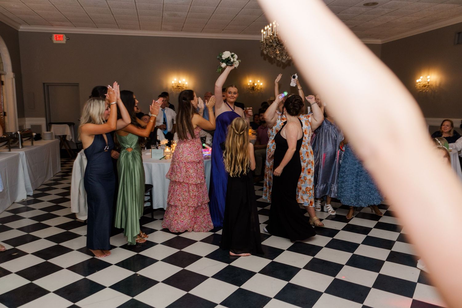 Wedding guests in colorful dresses gather on a black and white checkered dance floor during reception celebrations.