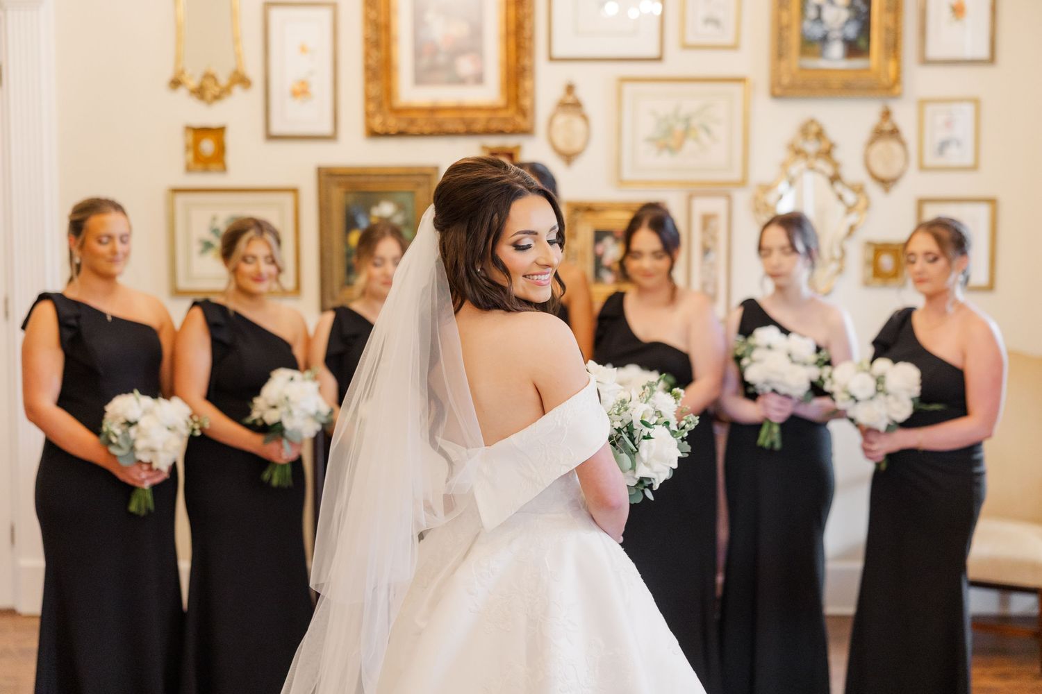 Wedding party members stand in line wearing black and white attire against an ornate indoor backdrop.