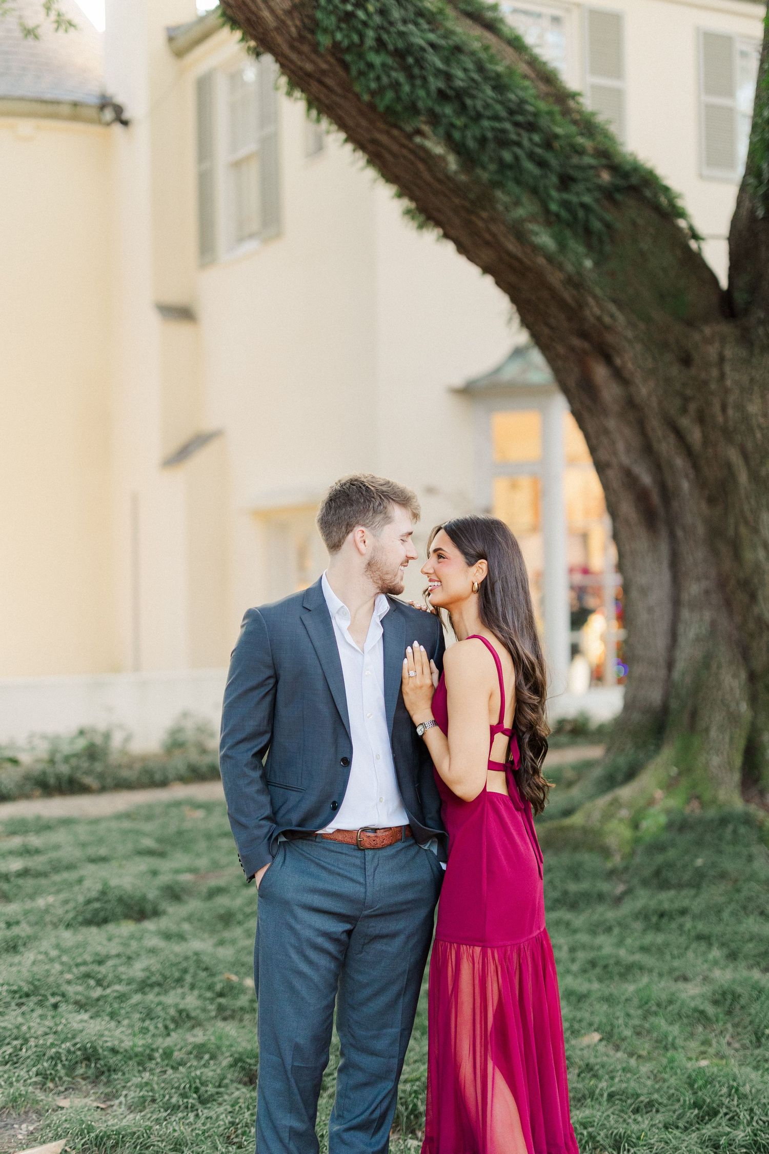 Romantic couple shares intimate moment under large tree with her in burgundy gown and him in dark suit.