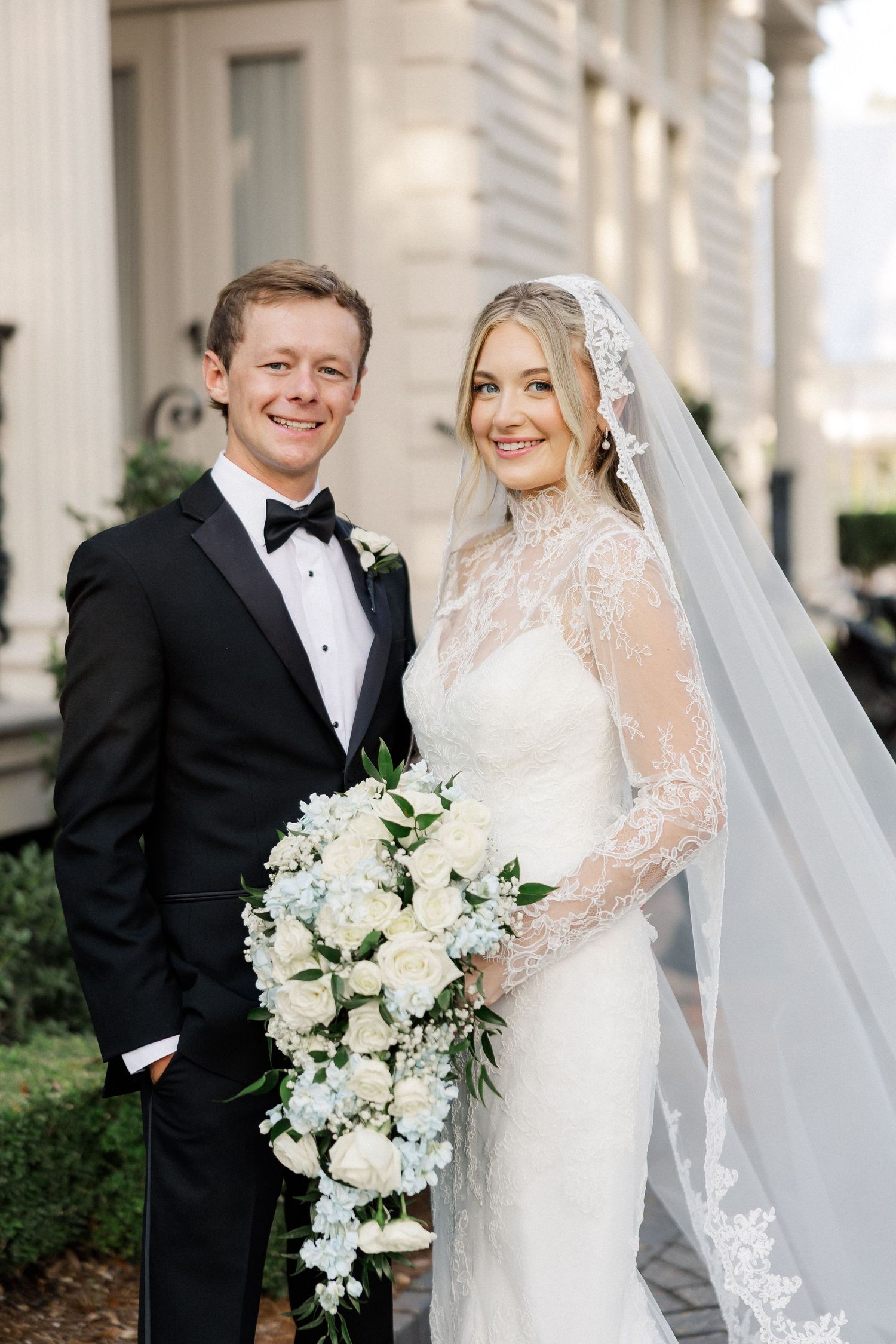 Elegant wedding couple poses in formal attire with white floral bouquet against classical architecture.