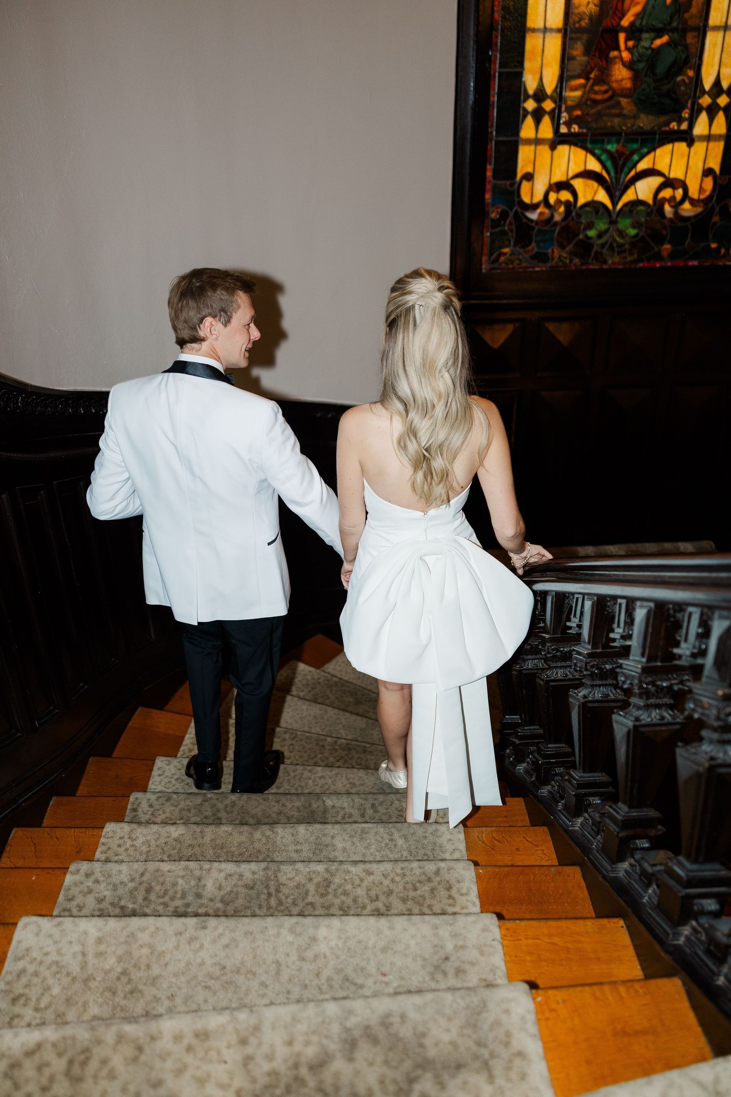 Couple ascending elegant staircase in formal wedding attire with stained glass window backdrop.