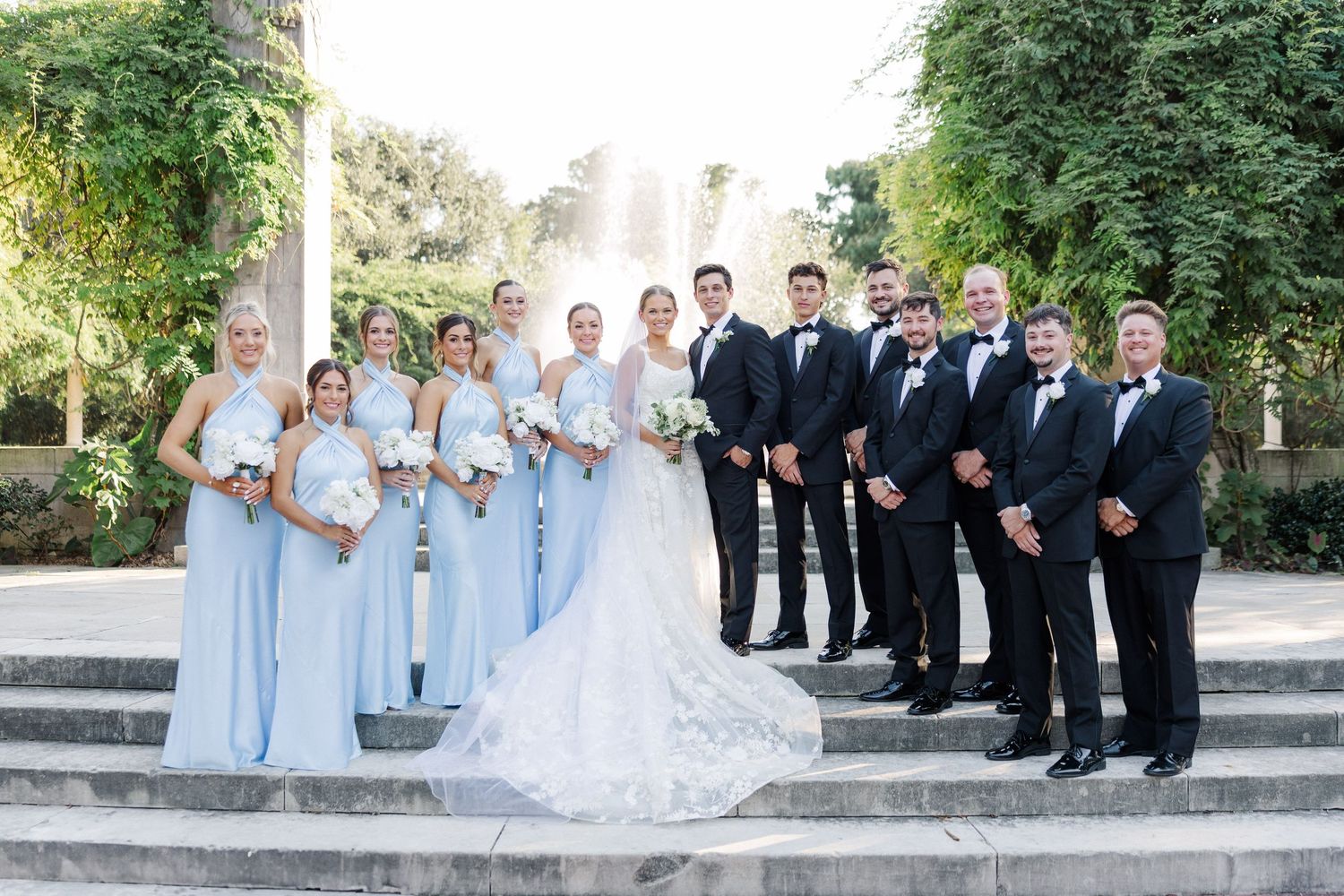 Wedding party in light blue dresses and black tuxedos posed on outdoor stone steps.