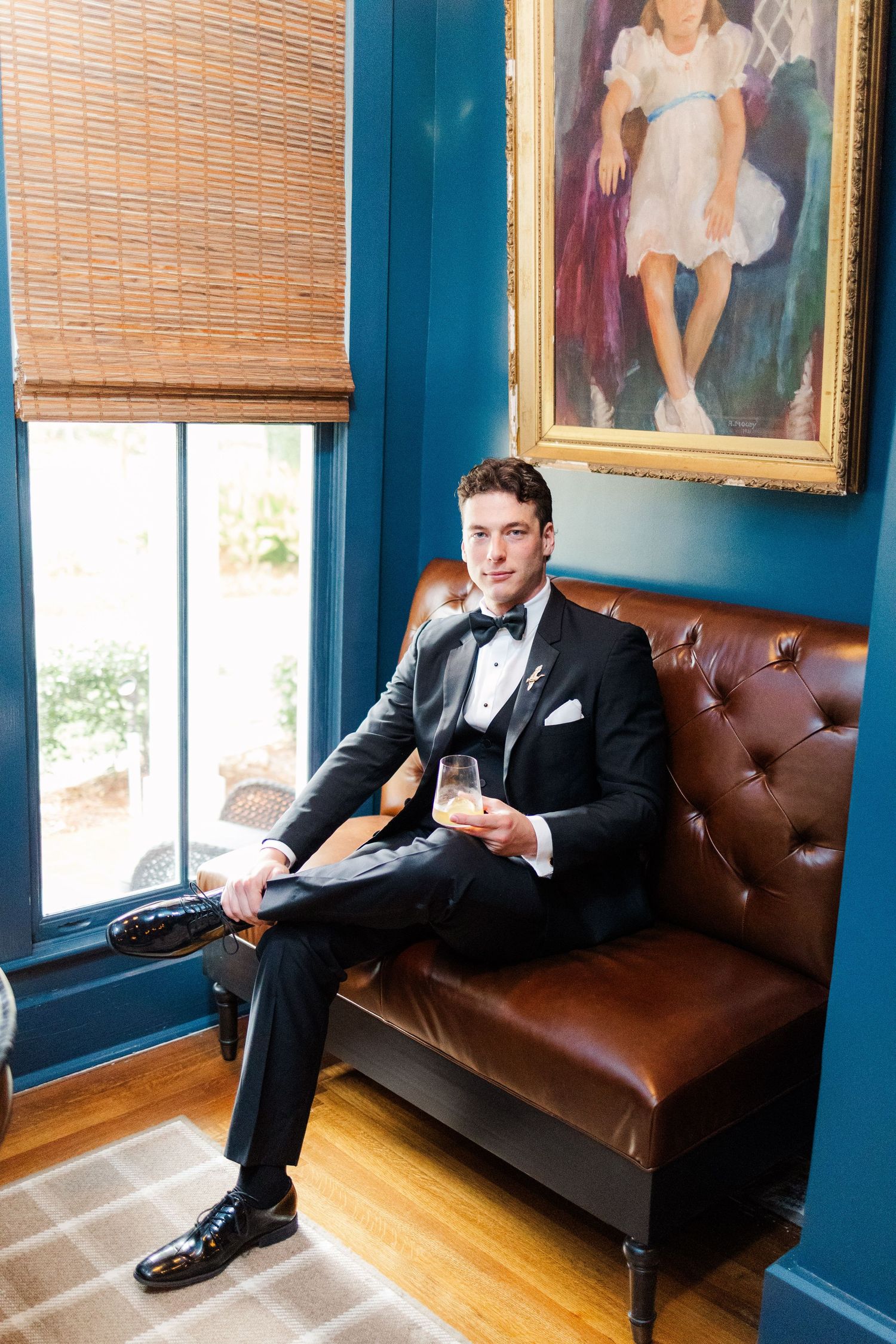 Groom in black tuxedo poses on brown leather couch against blue walls with portrait artwork.