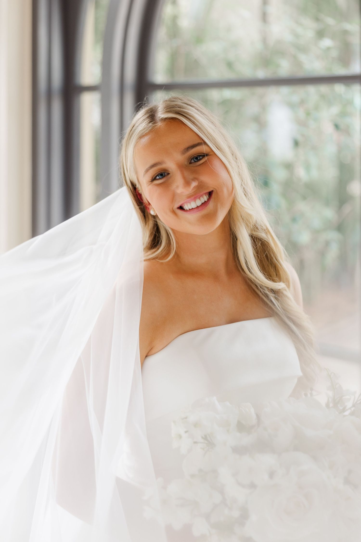 A flowing white wedding veil creates an ethereal effect in a bright sunlit room with large windows.