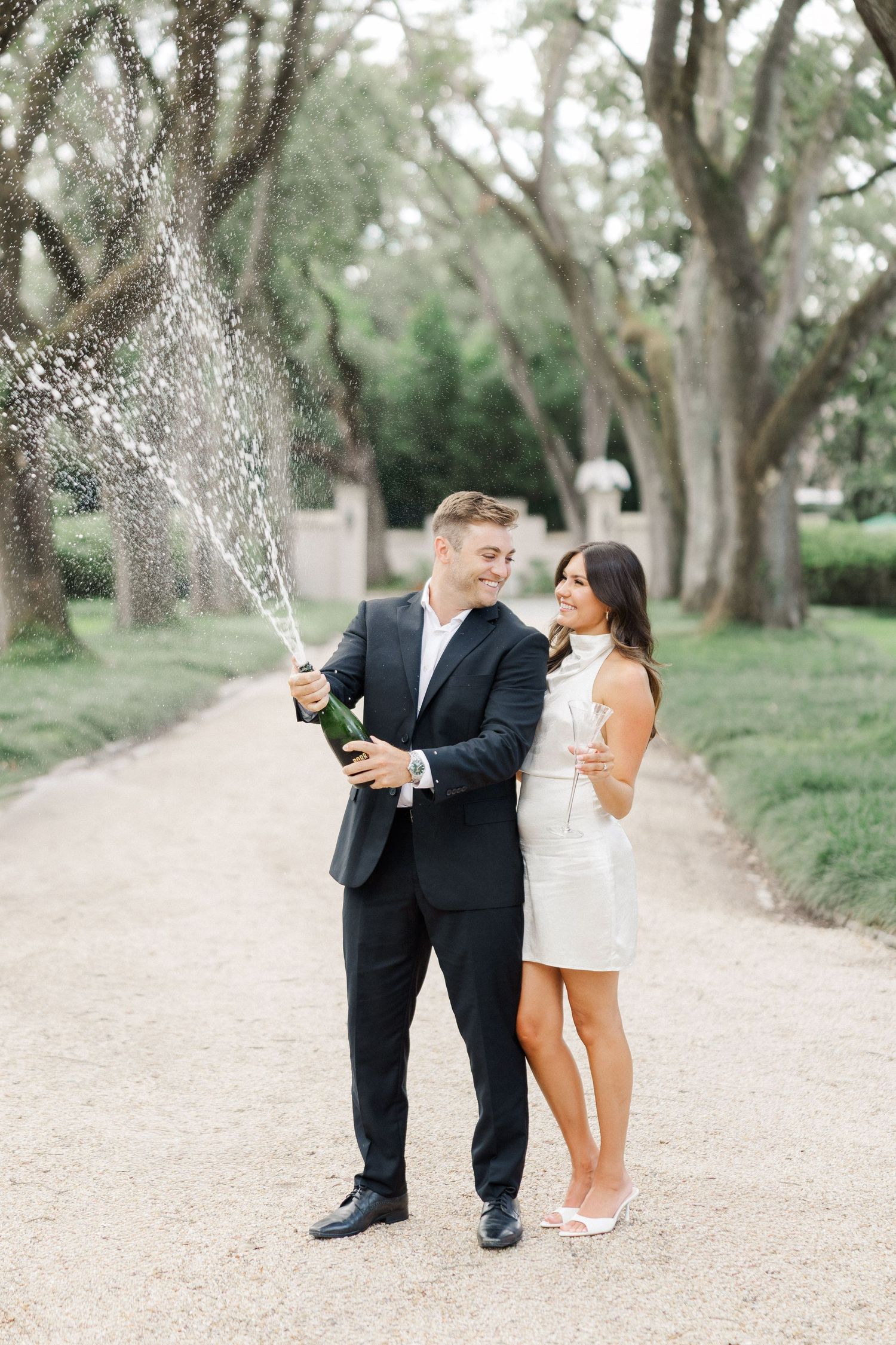 Couple celebrating engagement by popping champagne bottle on tree-lined garden path, man in black suit, woman in white mini dress.