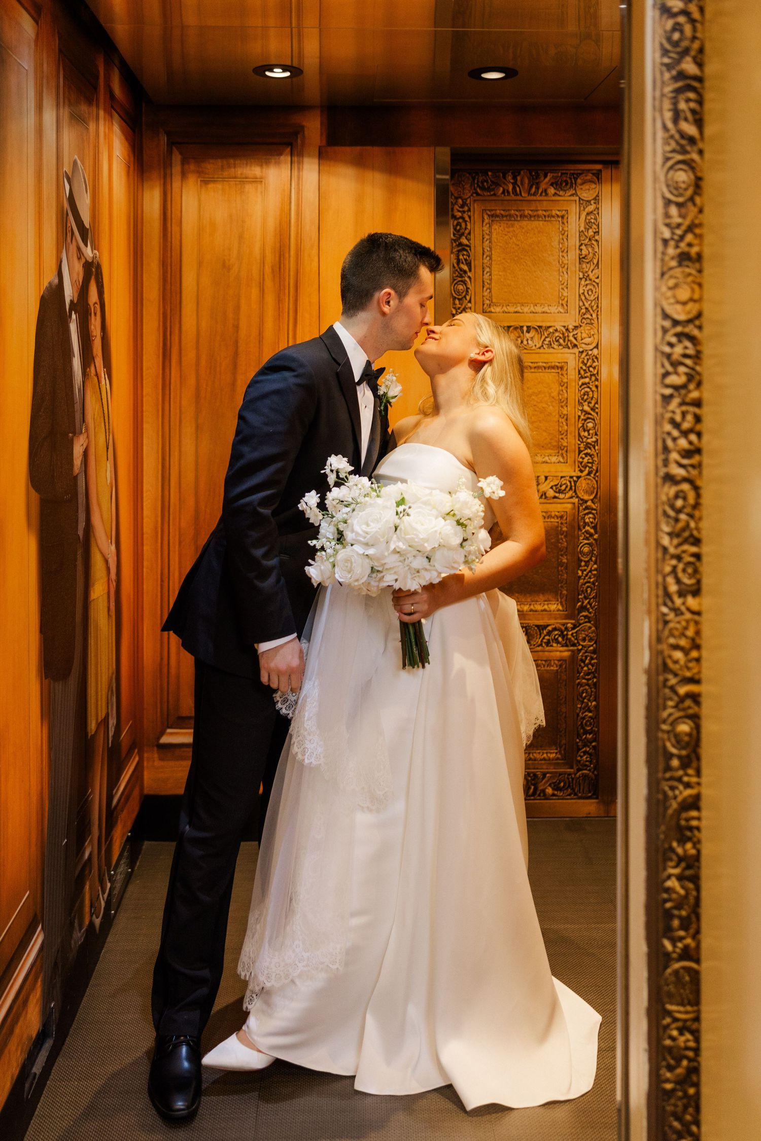 Elegant wedding couple shares a moment in wood-paneled hallway with ornate mirror.