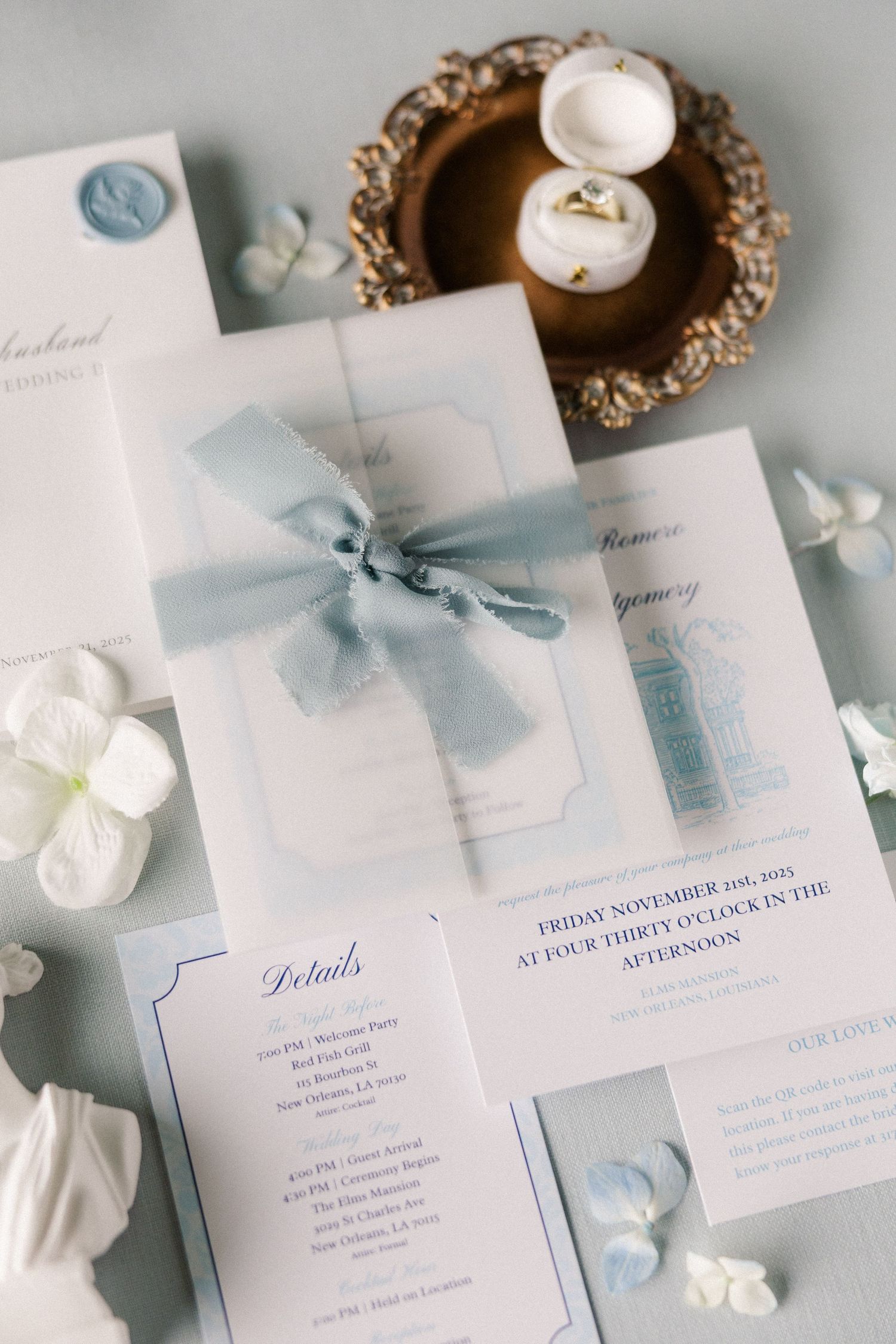 Elegant wedding invitation suite in white and pale blue with silk ribbons and wax seals laid out on light gray surface.
