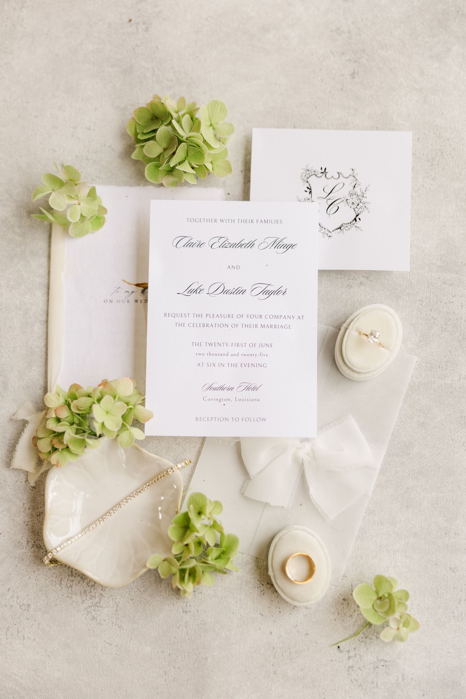 Elegant wedding invitation suite with green hydrangea flowers, white ribbons, and gold rings on light gray background.