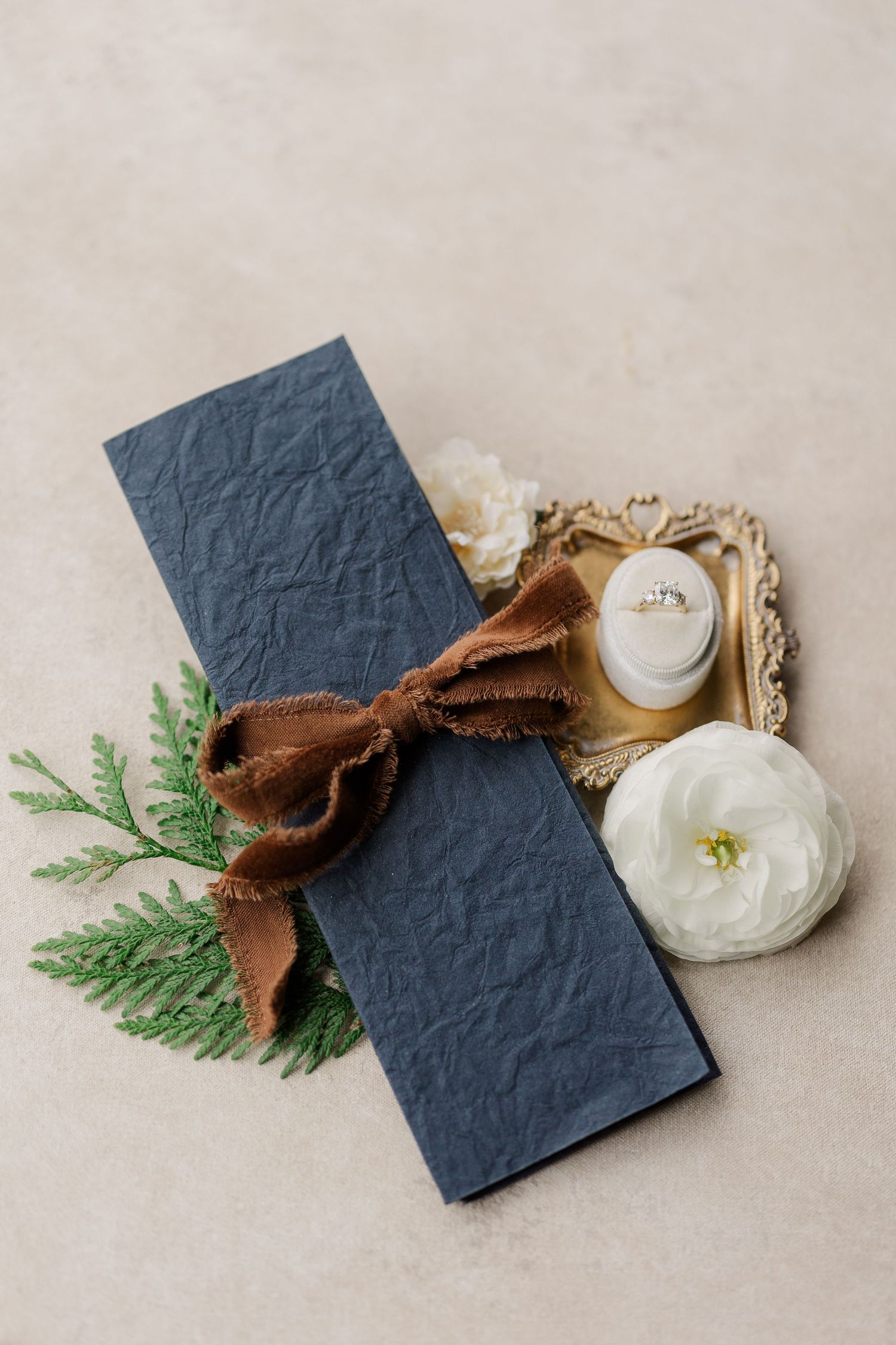 Elegant black invitation with brown velvet bow, evergreen sprig and white ranunculus flowers on beige background.