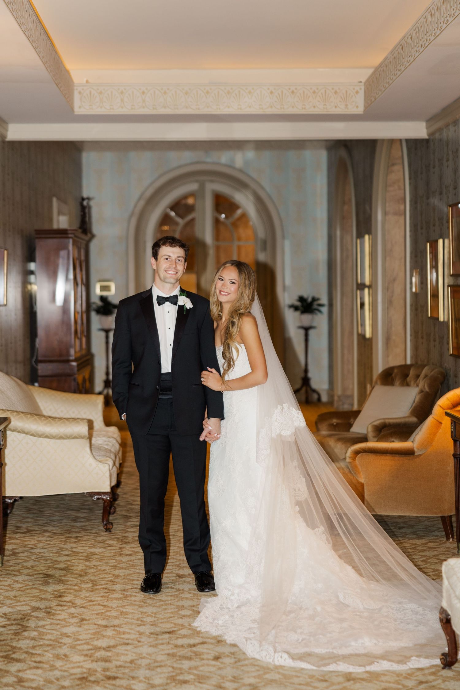 Elegant wedding couple walking through a luxurious hotel hallway with ornate carpet and decor.
