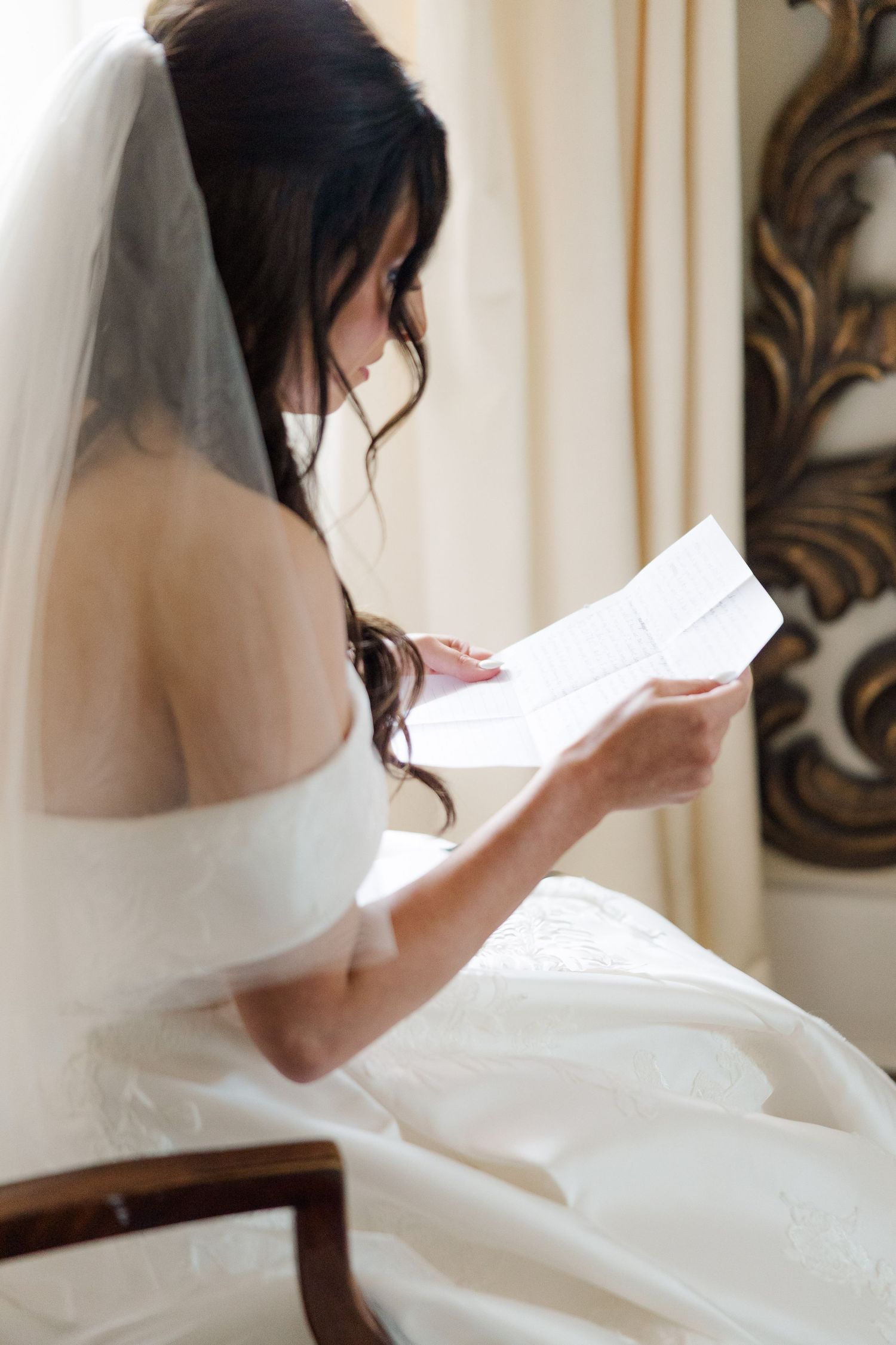 Person in white wedding dress reads a letter while sitting in elegant room.
