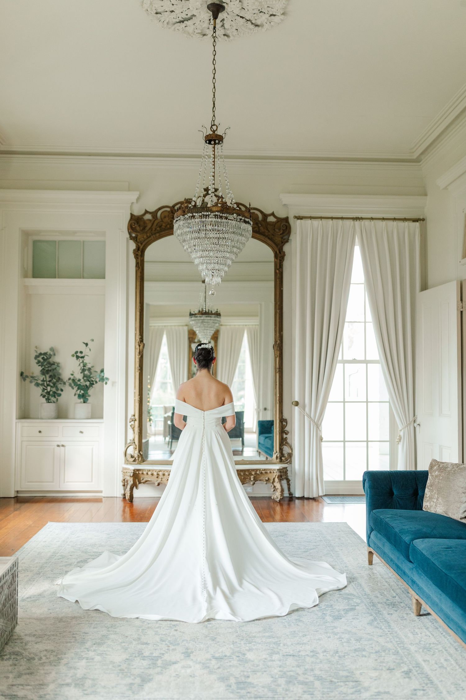 Elegant bridal gown with flowing train displayed in luxurious room with ornate gold mirror and crystal chandelier.
