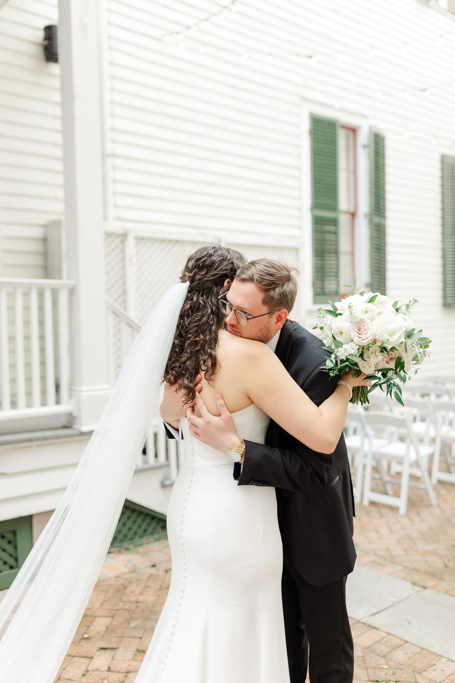A romantic wedding moment with couple embracing on a brick patio by a white house with green shutters.