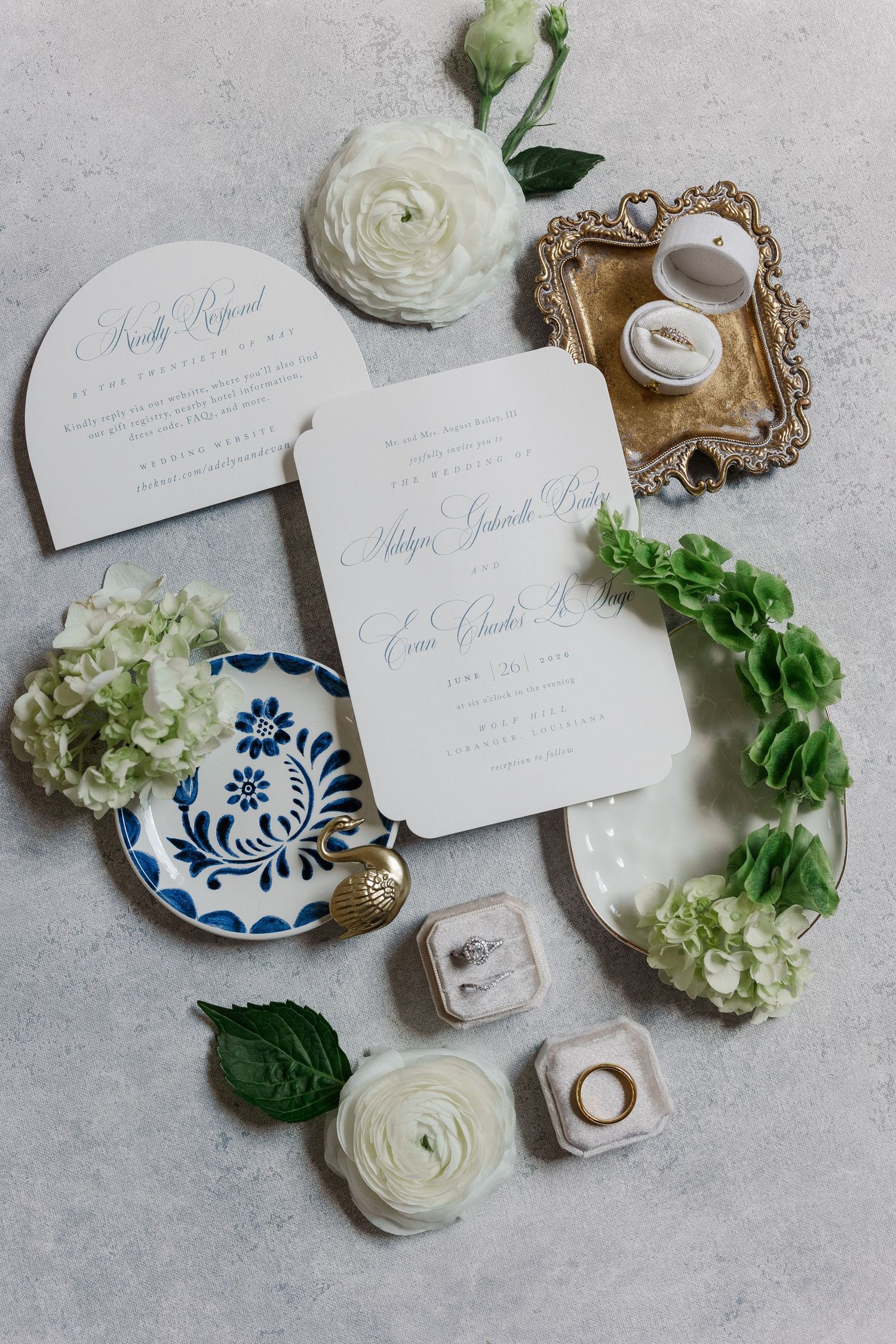 Elegant wedding invitation suite with white florals, blue ceramic dish, gold tray, and ring boxes on gray surface.