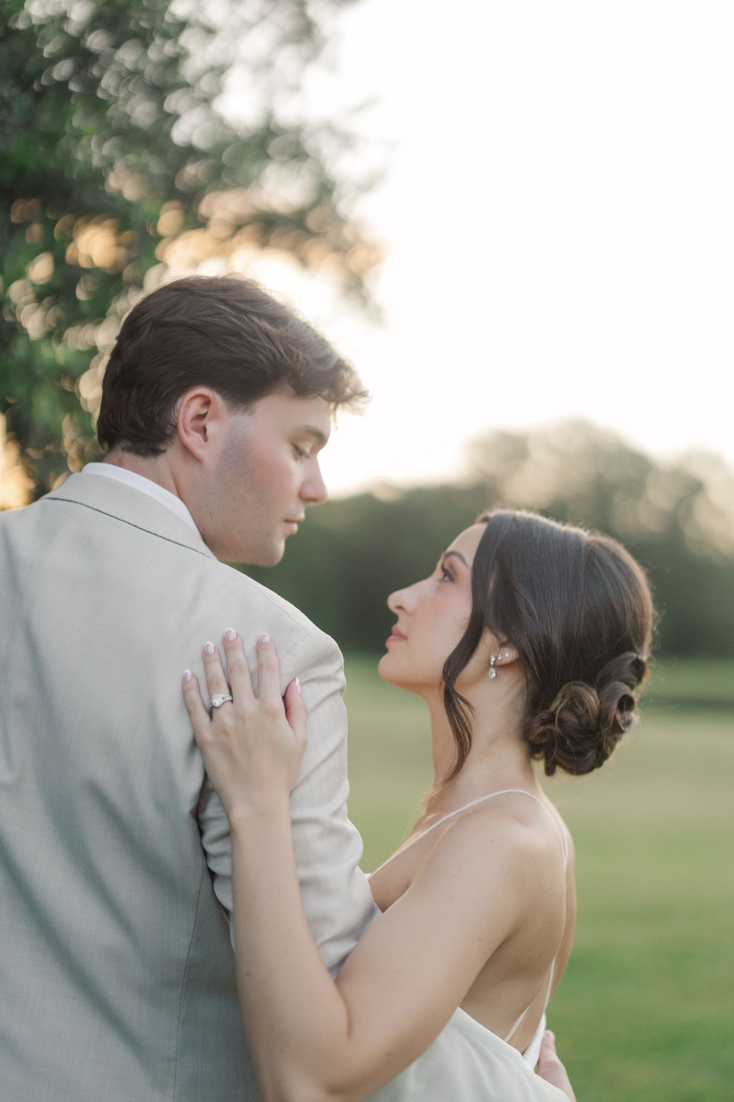 Bride and groom share a tender moment at sunset, nearly kissing, bride in white gown, groom in beige suit.