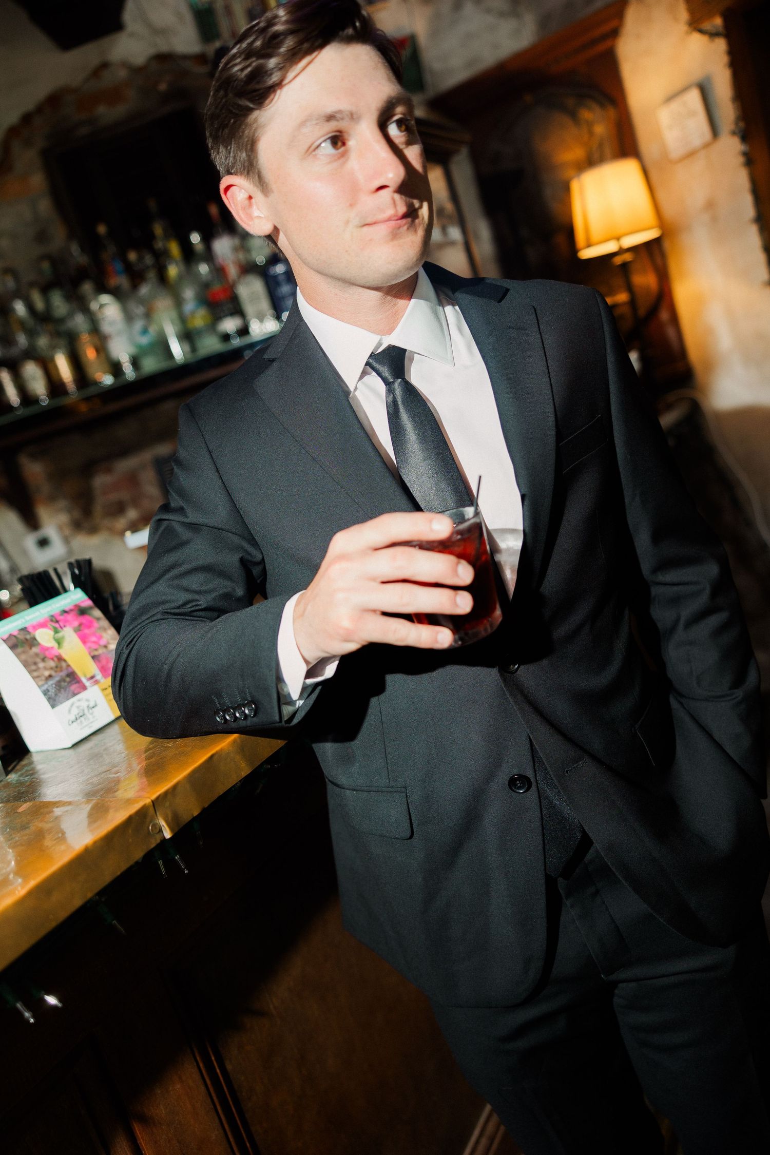 Dapper groom in dark suit holding cocktail while leaning against a bar at wedding reception.
