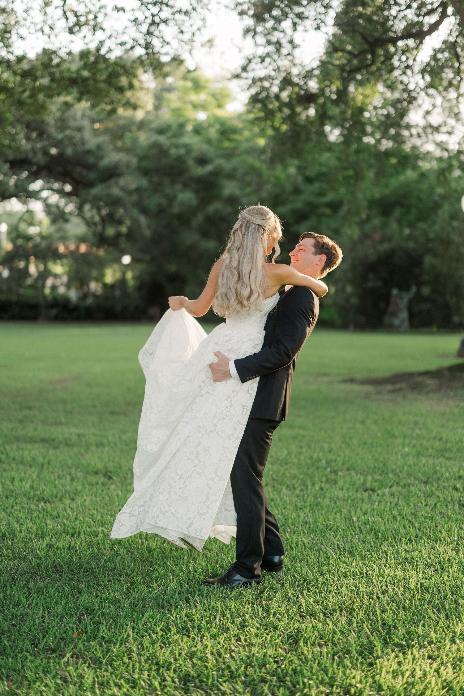 Groom in black suit lifts bride in white lace gown on green lawn during outdoor wedding photos.