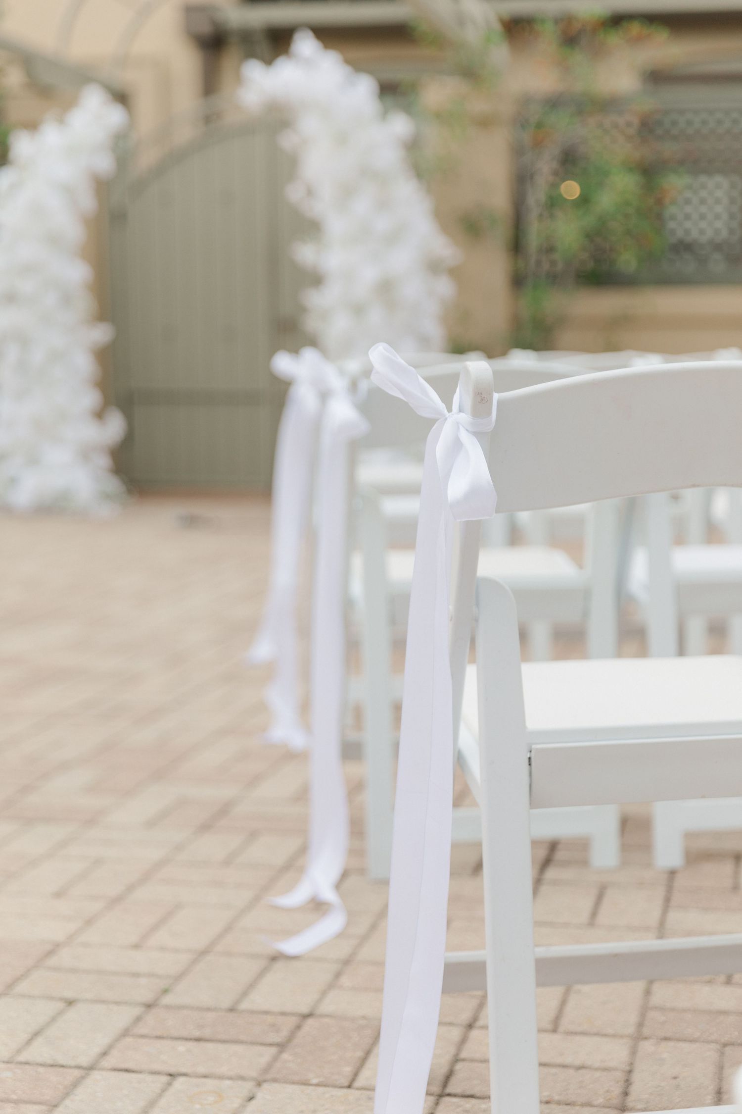 White folding chairs with decorative bows line a stone patio for an outdoor wedding ceremony.