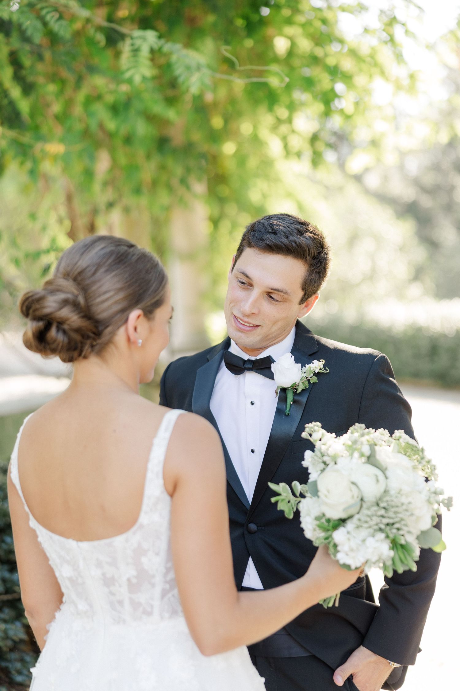 A bride and groom share a tender moment outdoors surrounded by greenery and white flowers.