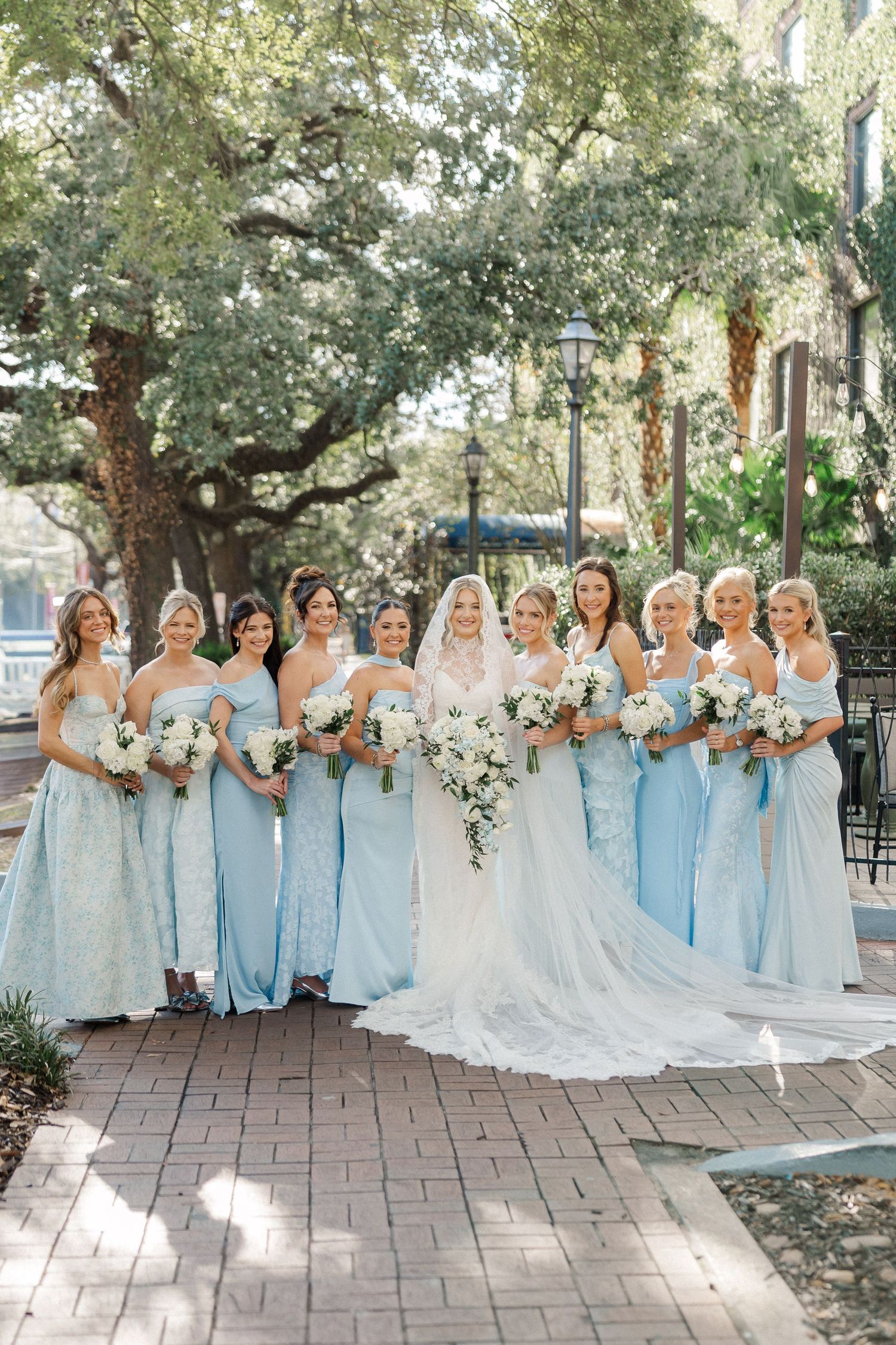 Wedding party in light blue bridesmaid dresses and white bridal gown pose outdoors on brick patio.
