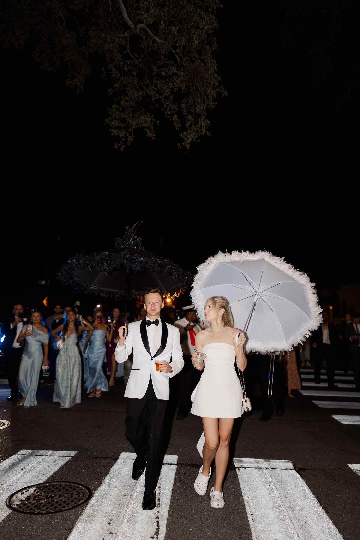 A couple with white parasols walks along a lit pathway during their nighttime wedding exit.
