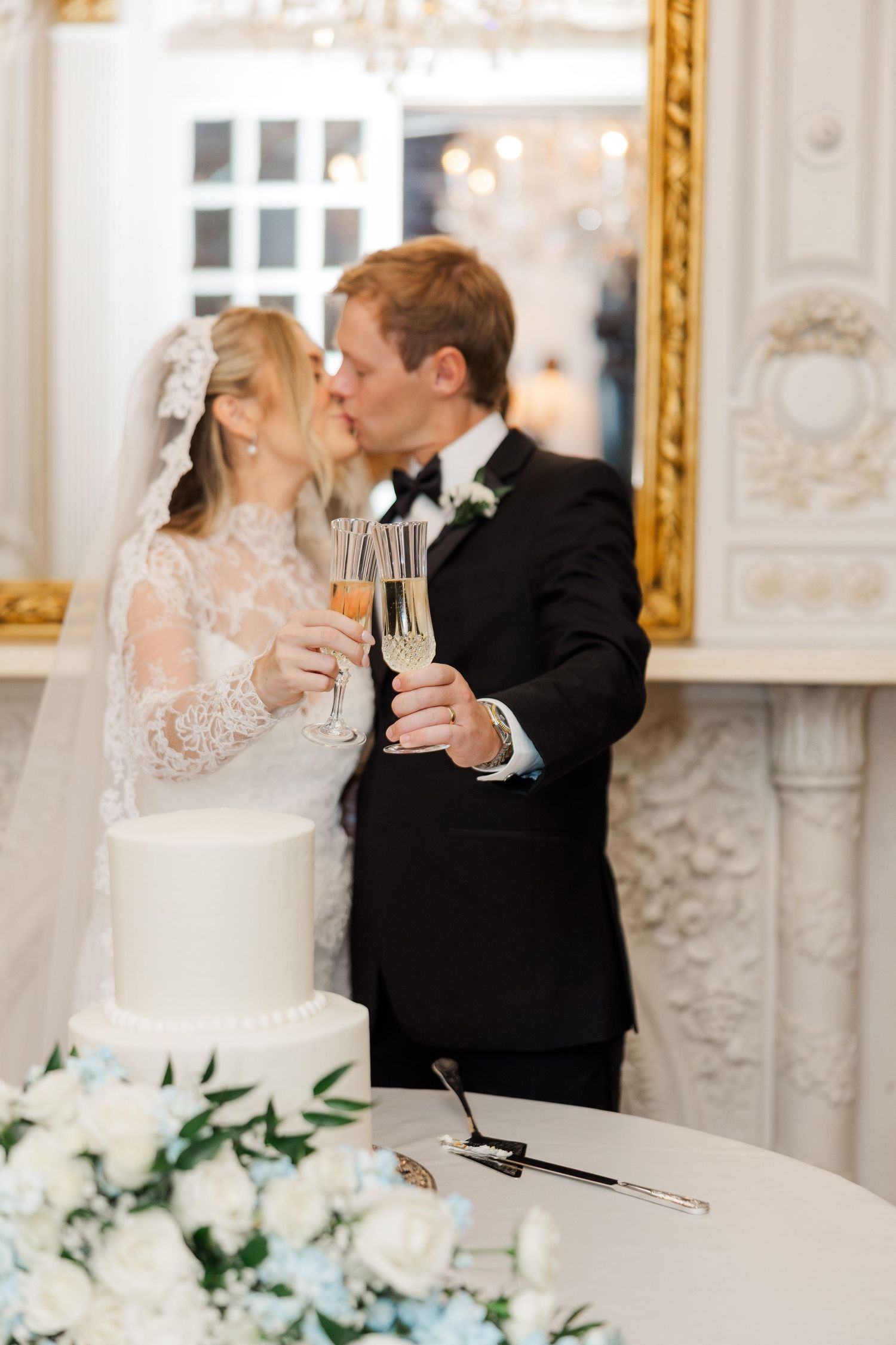 A wedding couple shares a champagne toast in an ornate white and gold reception room.