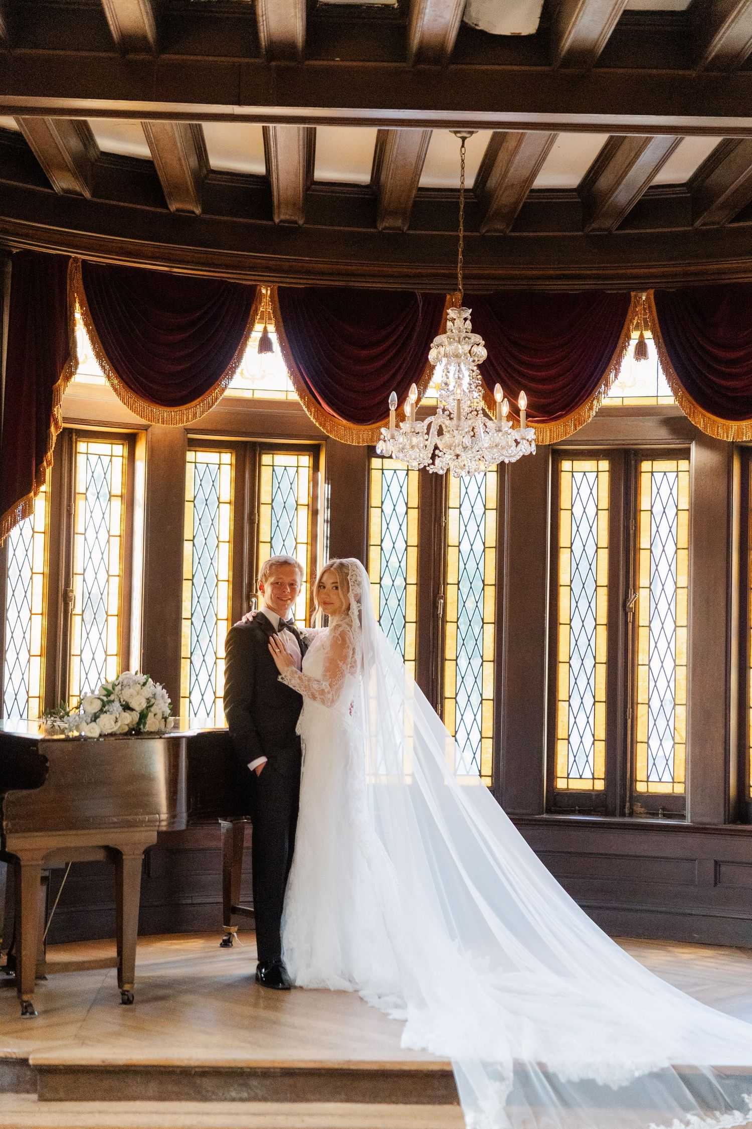 An elegant ballroom with wood paneling features stained glass windows and a crystal chandelier.