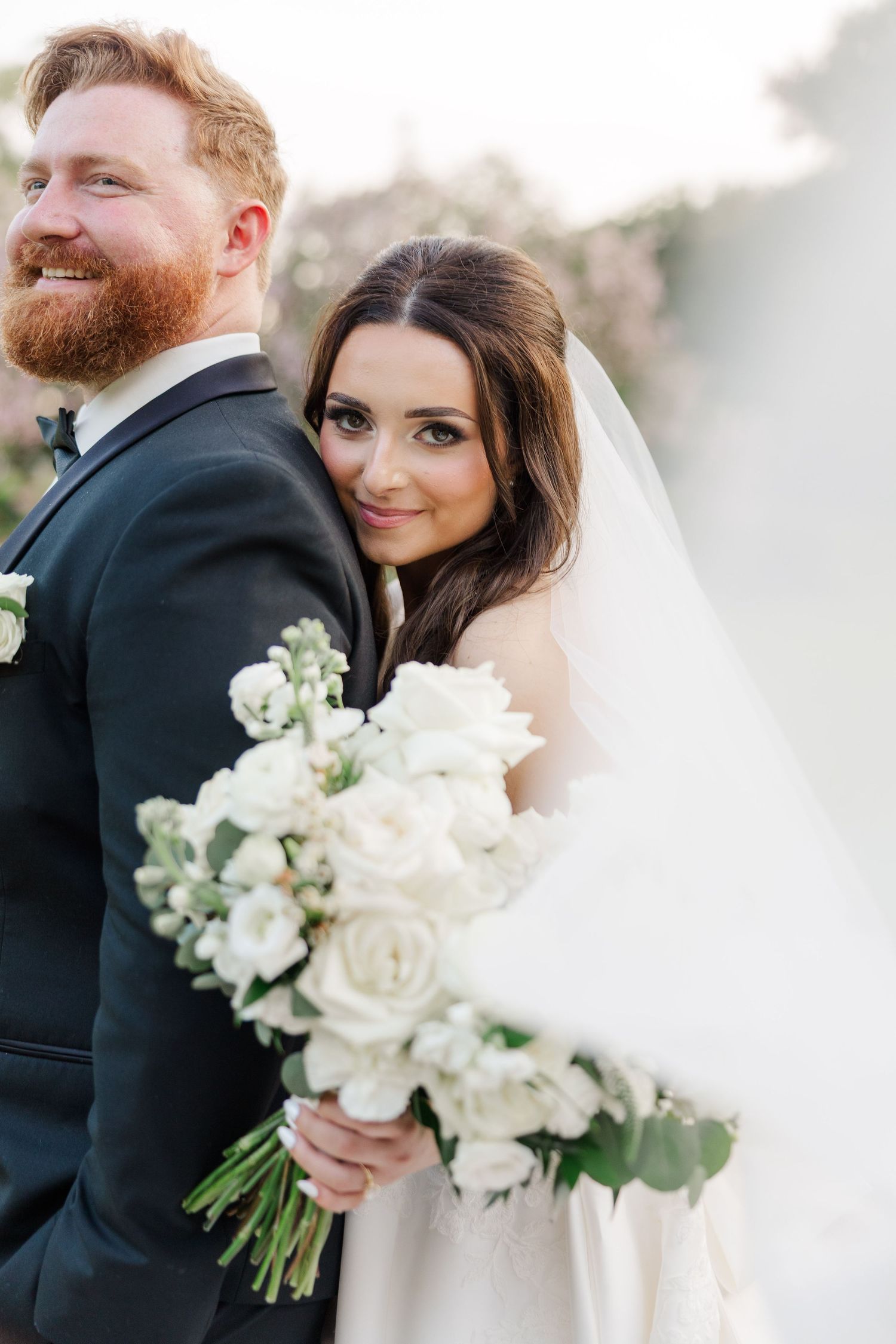 Elegant wedding couple poses with white floral bouquet against soft background.