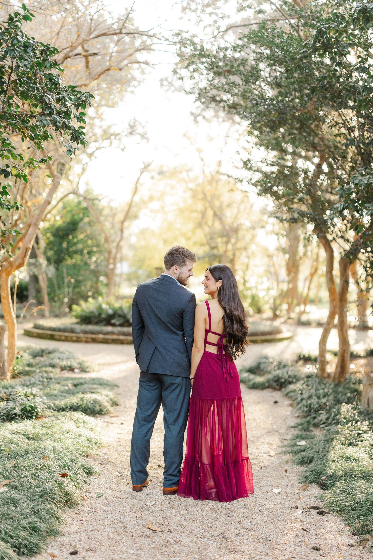 Couple walks down sunlit garden path surrounded by trees and greenery.