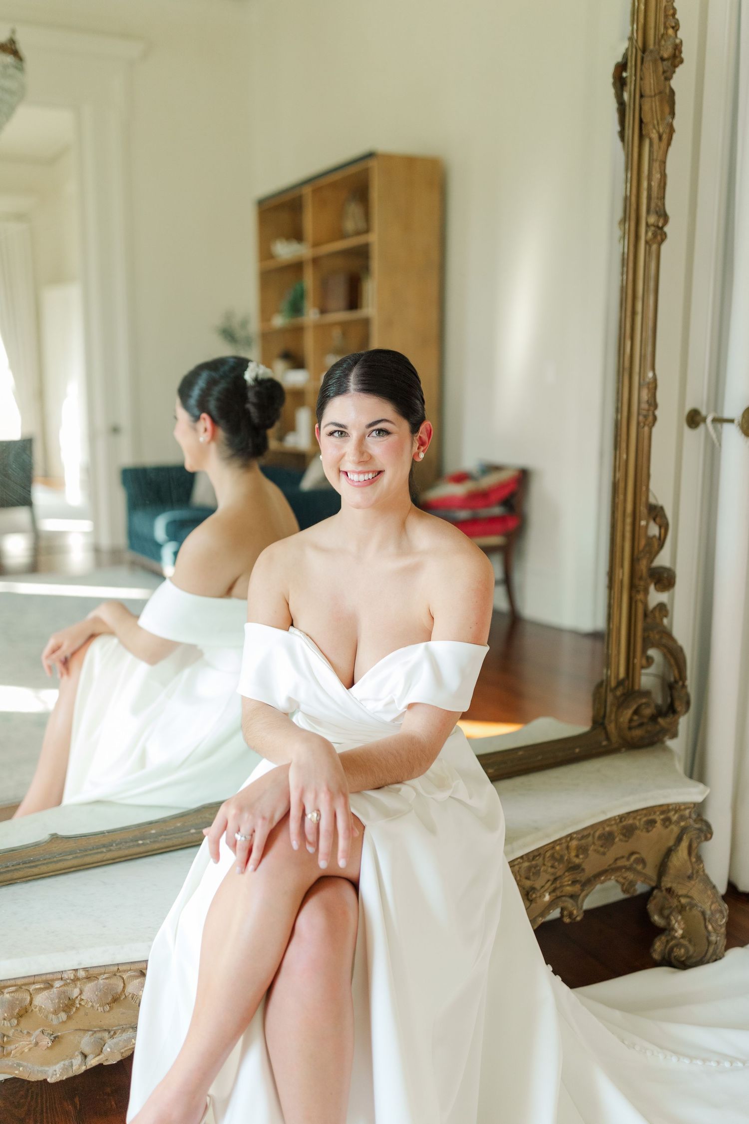 A bride in a white off-shoulder wedding dress sits elegantly on an ornate mirror bench in natural light.