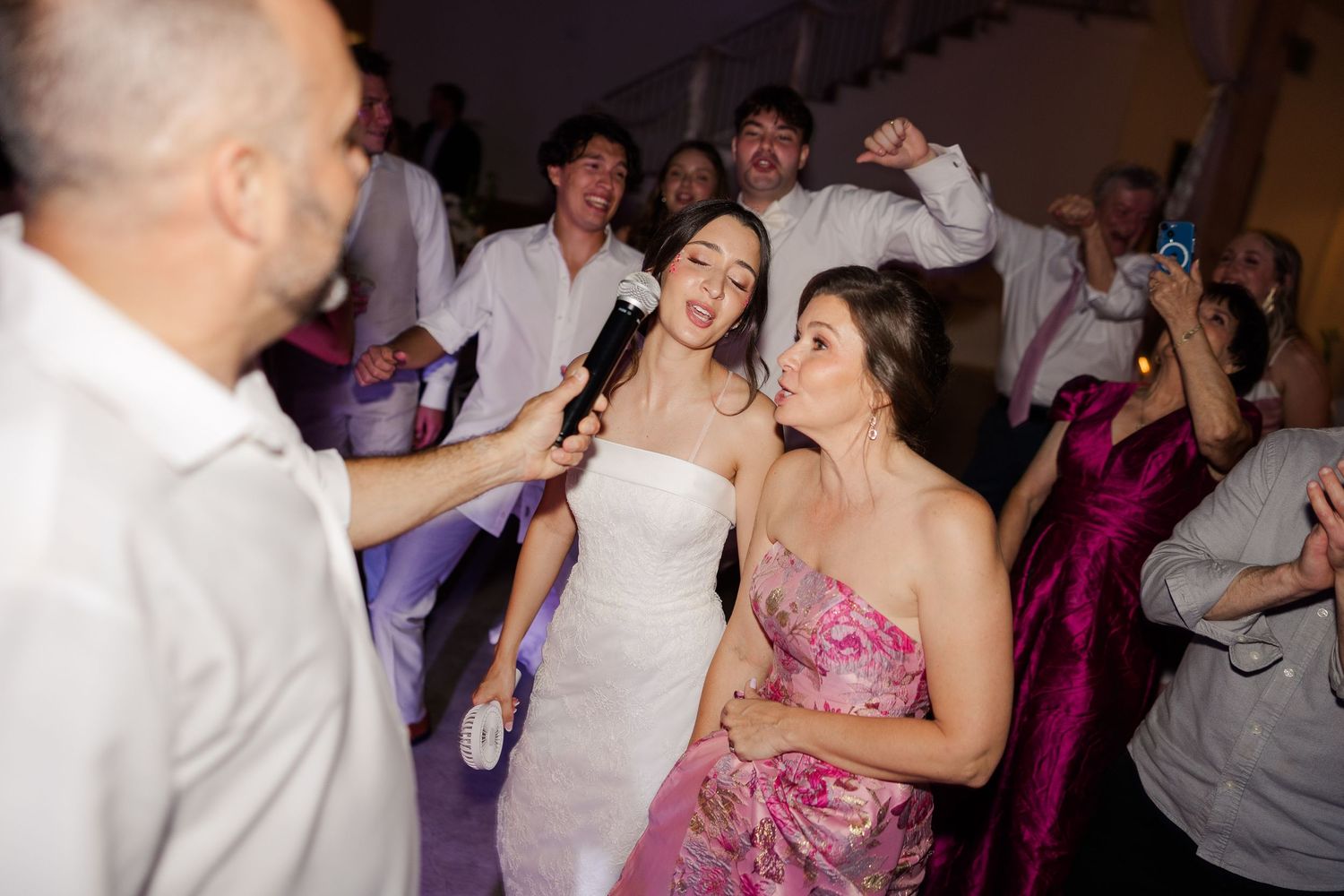 Joyful bride in white strapless gown sings into microphone with friend in floral dress on crowded dance floor.