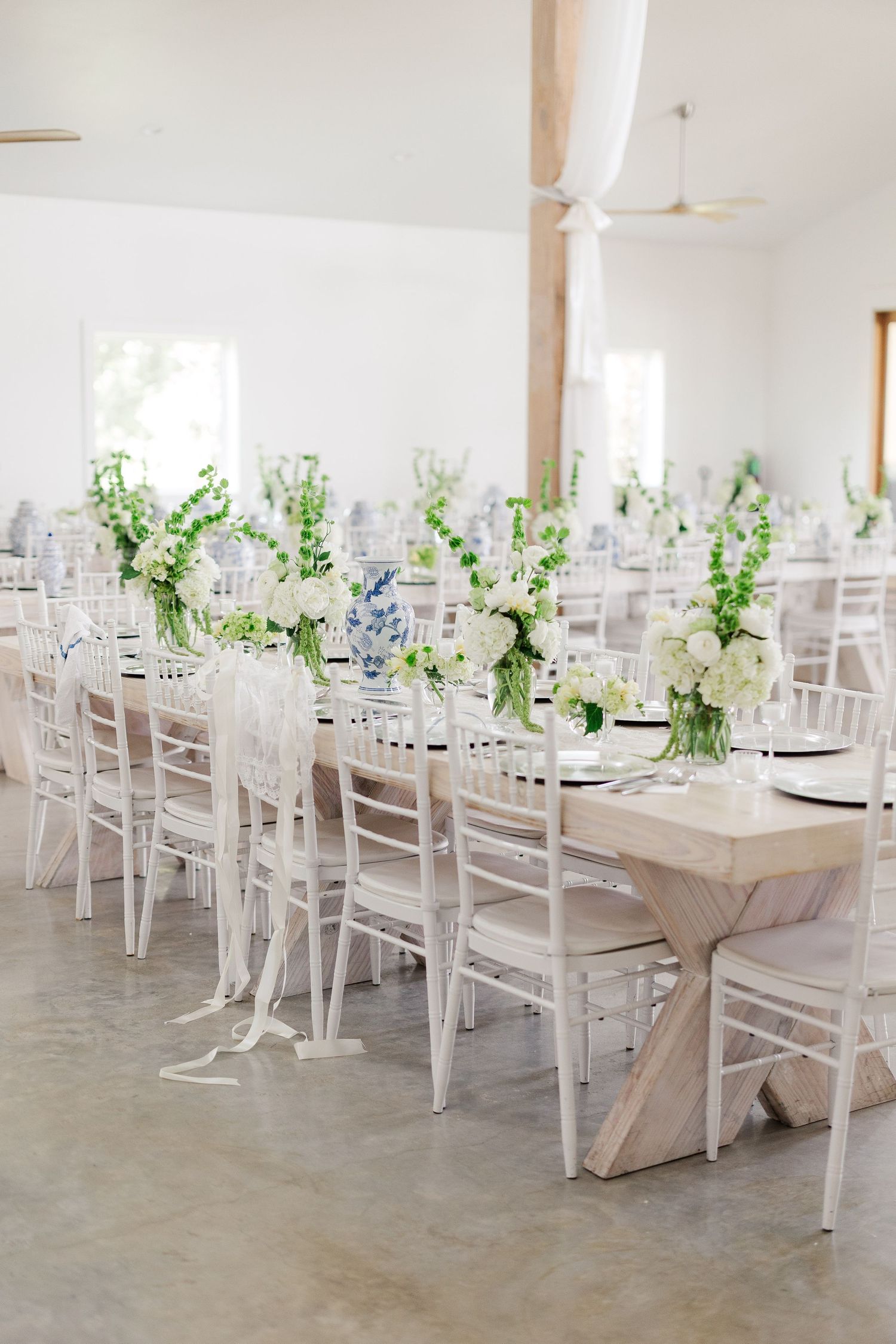 Elegant white barn wedding reception with long farm tables, chiavari chairs, and white floral centerpieces.