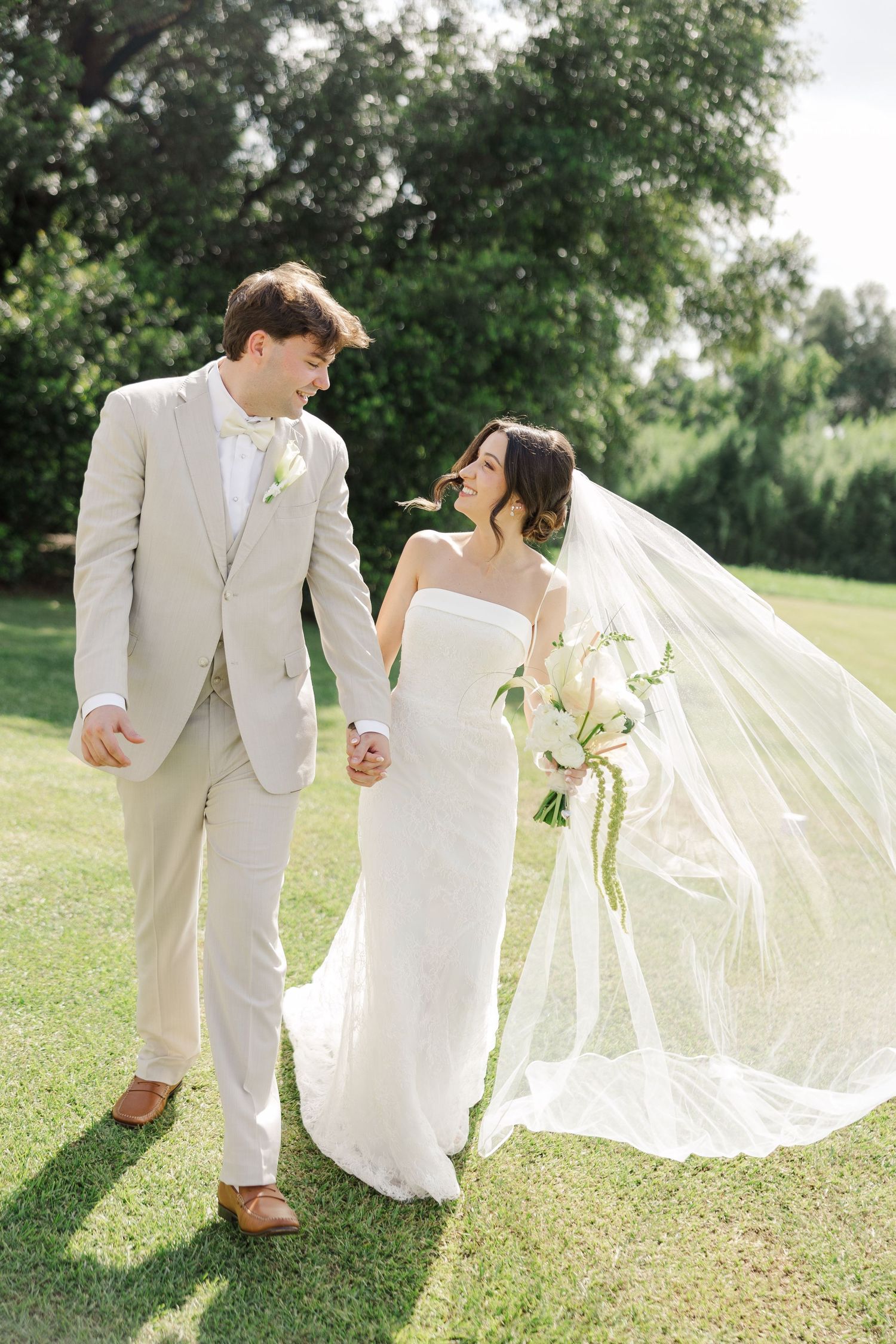 Smiling bride and groom walk hand-in-hand on green lawn, her veil flowing in the breeze, holding bouquet.
