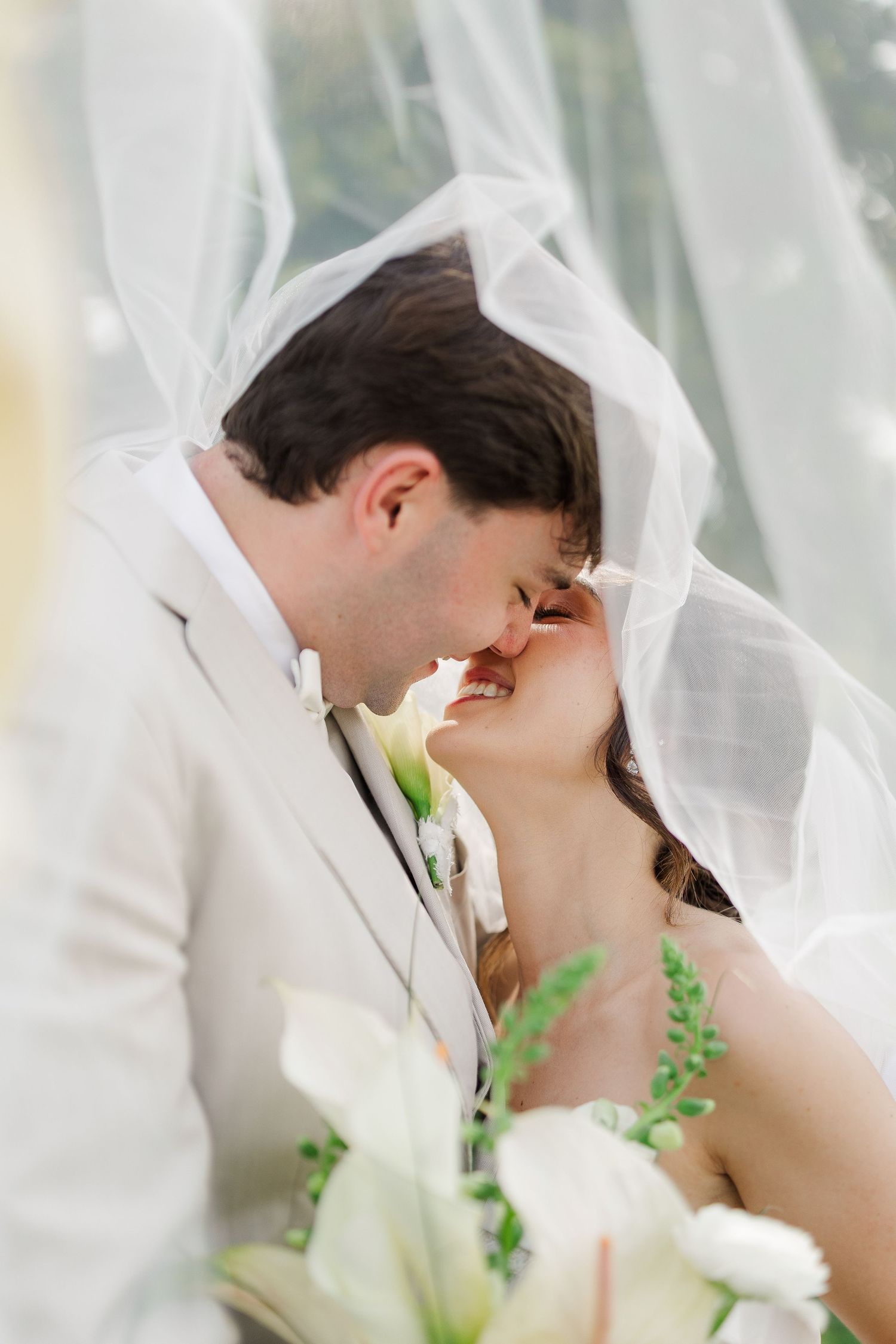 Bride and groom share a romantic kiss under flowing white veil, with white floral bouquet in foreground.
